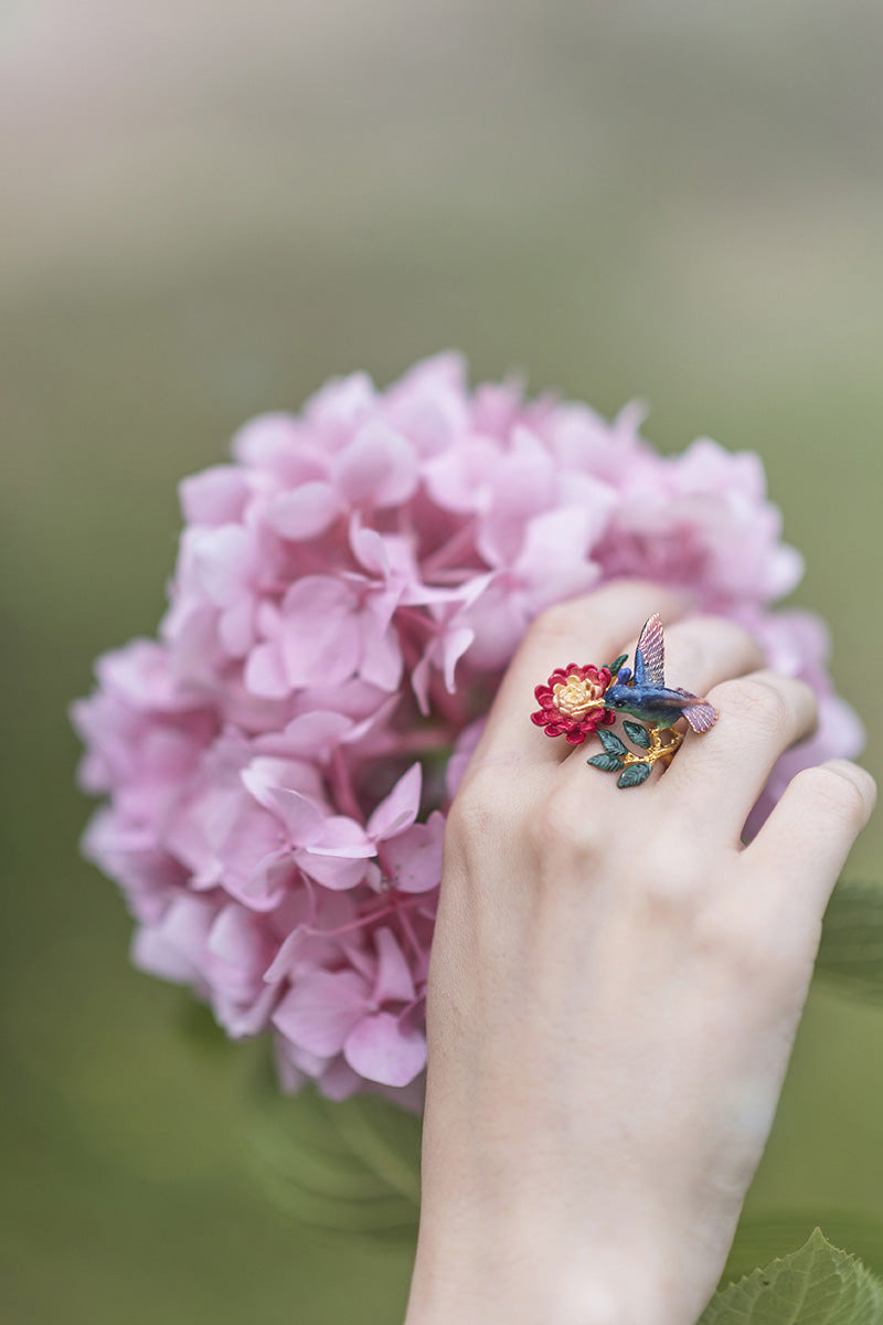 A hand adorned with a colorful hummingbird ring, surrounded by lush pink hydrangeas.