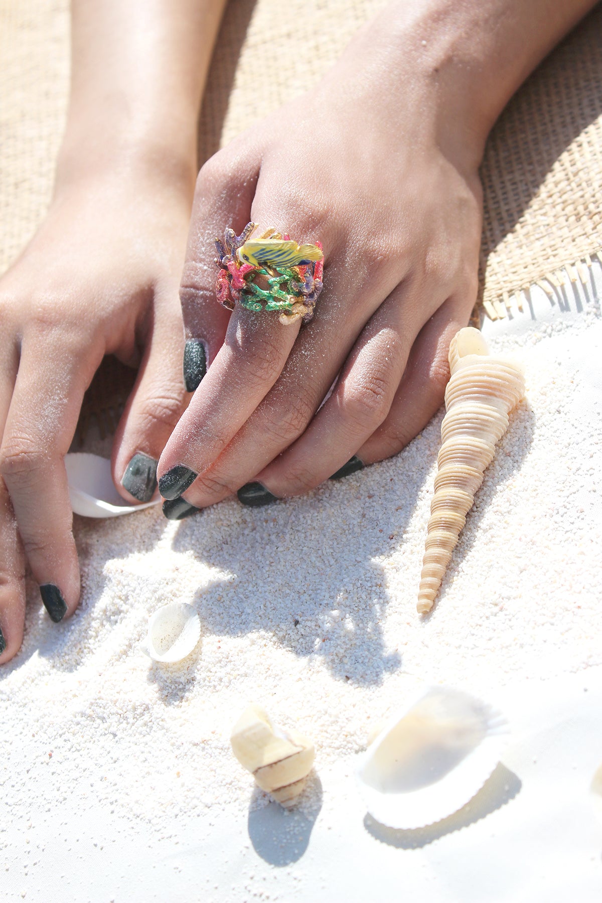 A close-up of a hand wearing a colorful fish with coral reefs ring on a sandy surface.