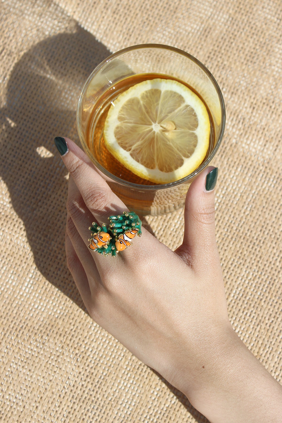 A hand wearing a colorful clownfish ring holding a glass of lemon drink.