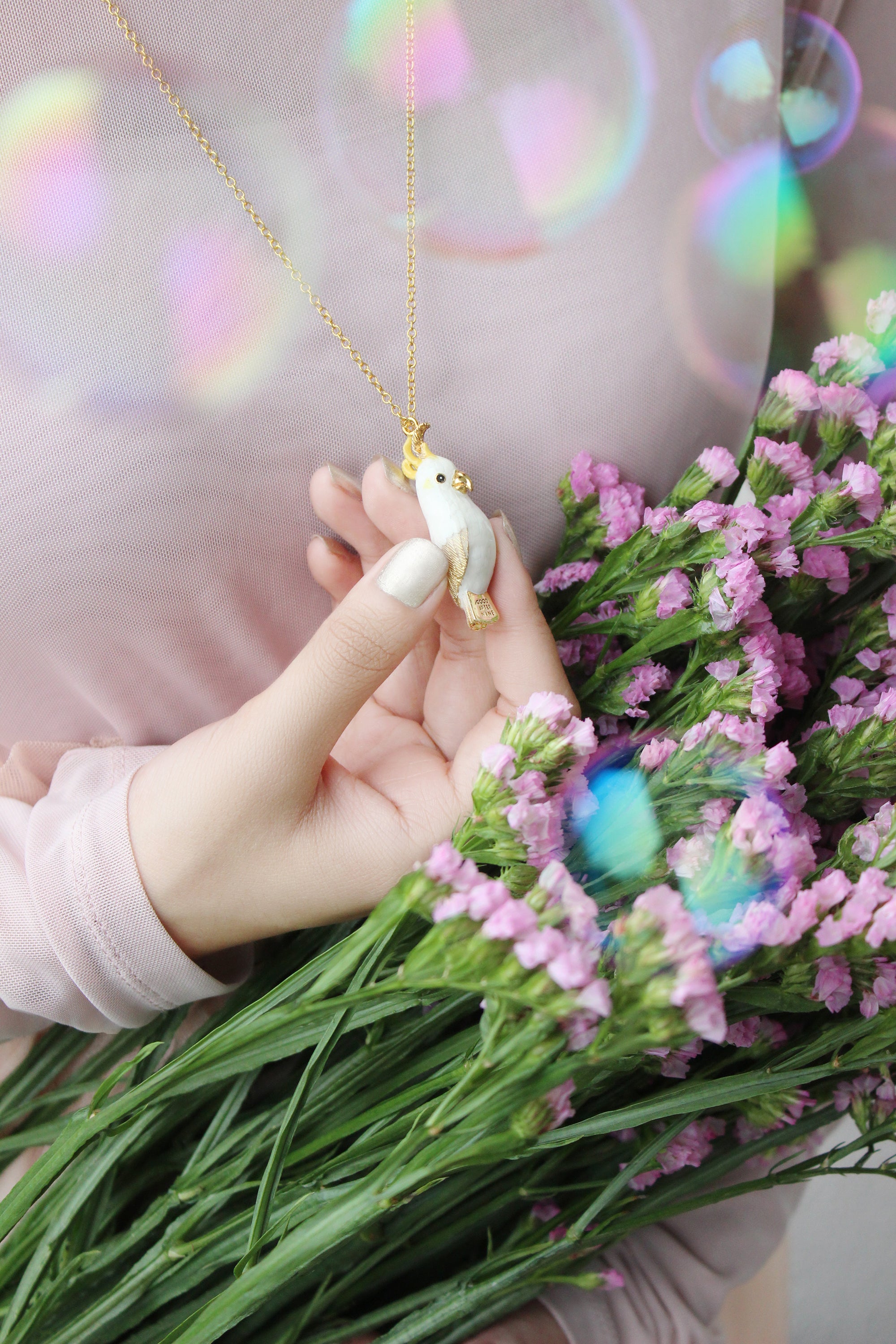 Cockatoo pendant necklace held by a hand, with a bouquet of pink flowers in the foreground.