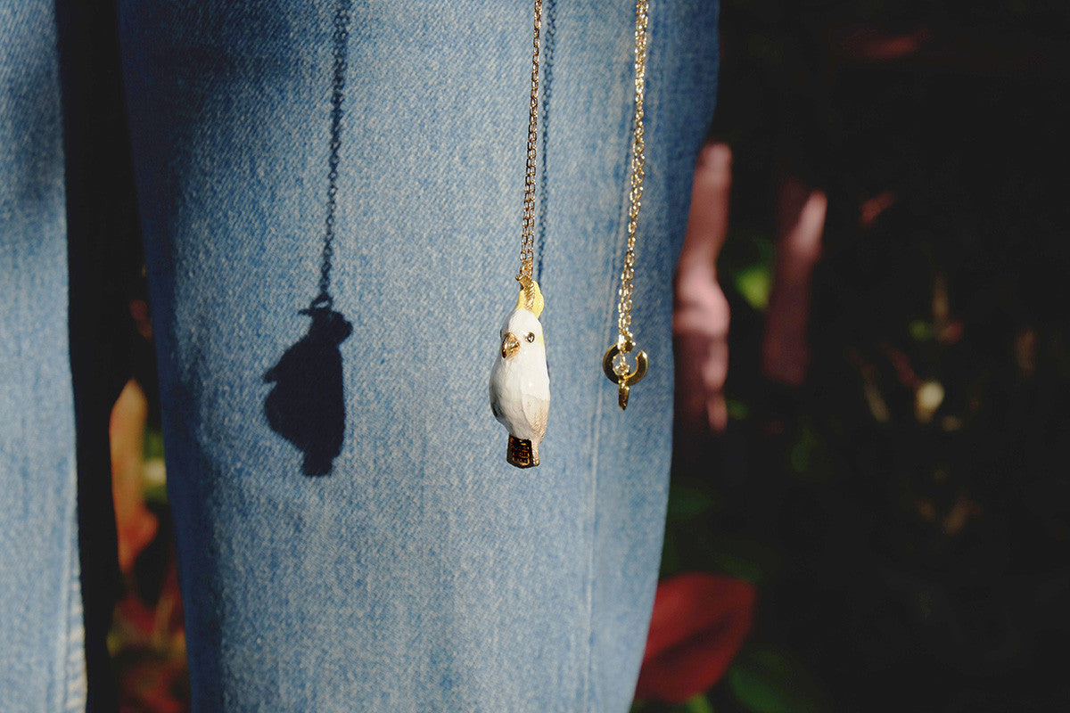 Gold chain necklace featuring a cockatoo whistle pendant hanging beside the shadow on denim fabric.