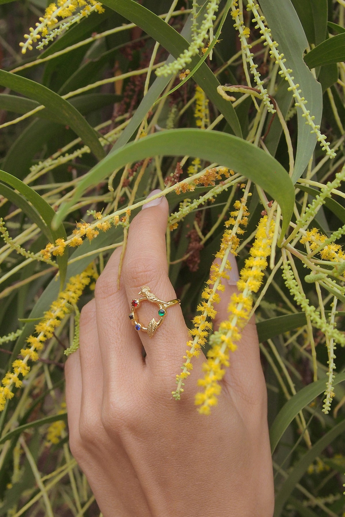 A hand wearing a delicate gold ring featuring a bird design and various colored stones among vibrant yellow flowers and green leaves.
