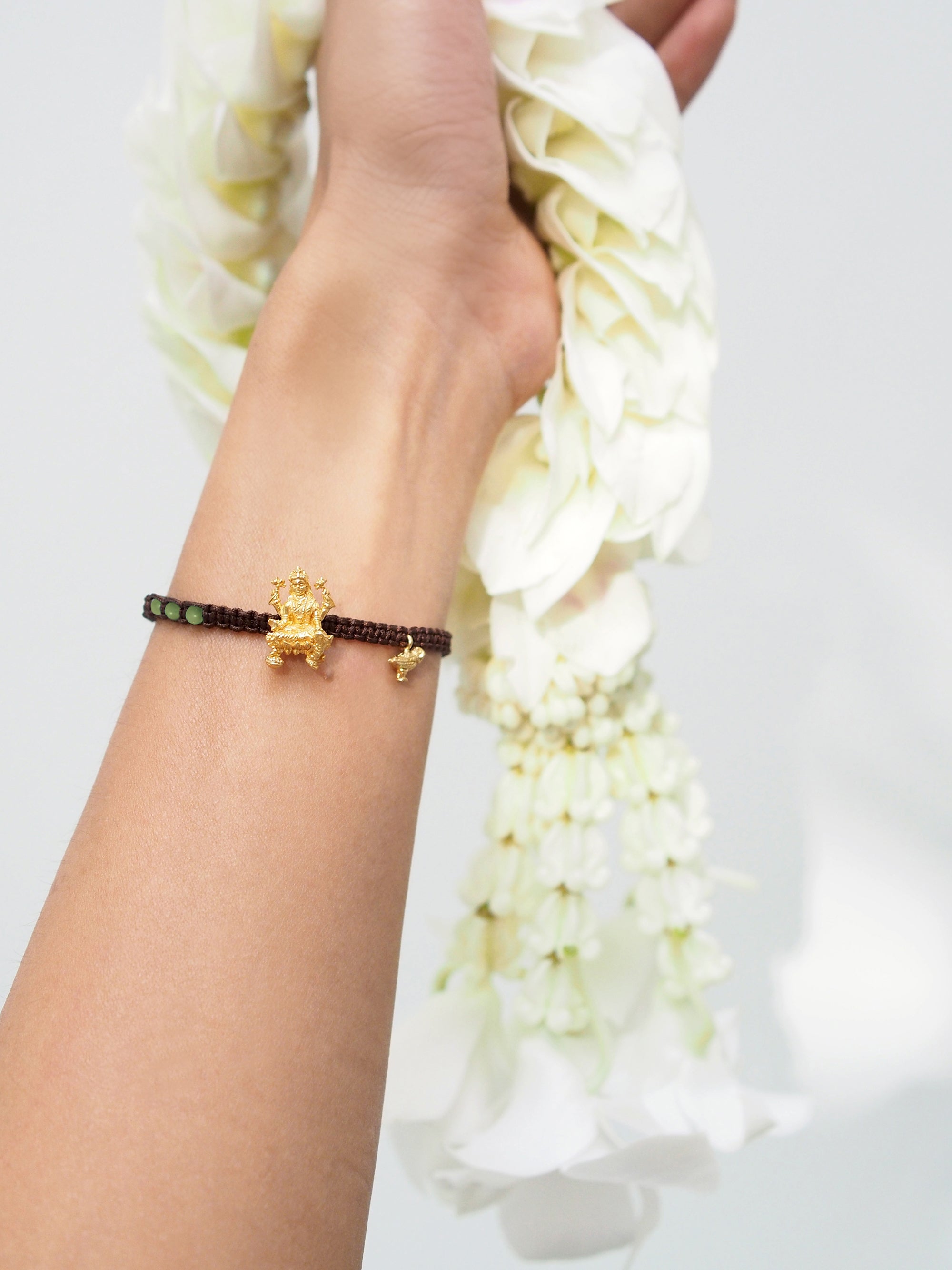 Close-up of a hand wearing a brown bracelet featuring Tridevi Lakshmi and owl charms, while holding a garland.