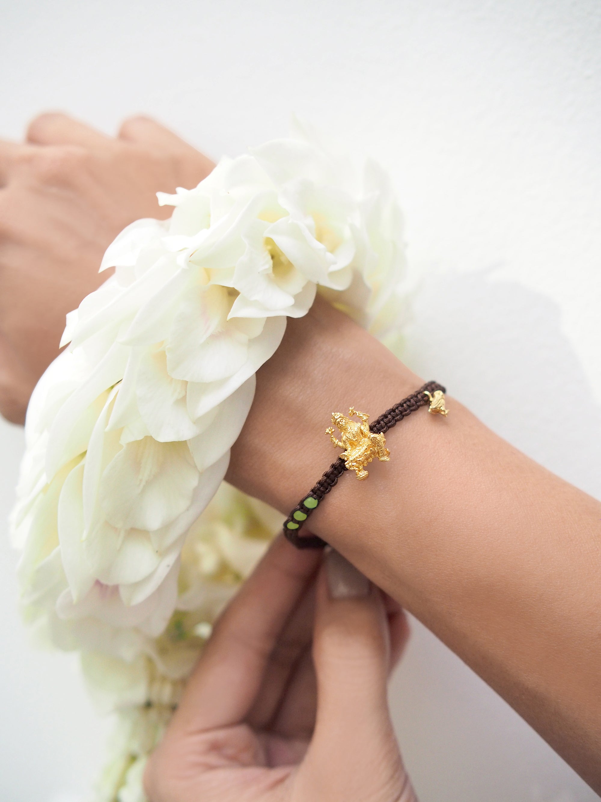 Close-up of a hand wearing a brown bracelet featuring Tridevi Lakshmi and owl charms, while holding a garland.