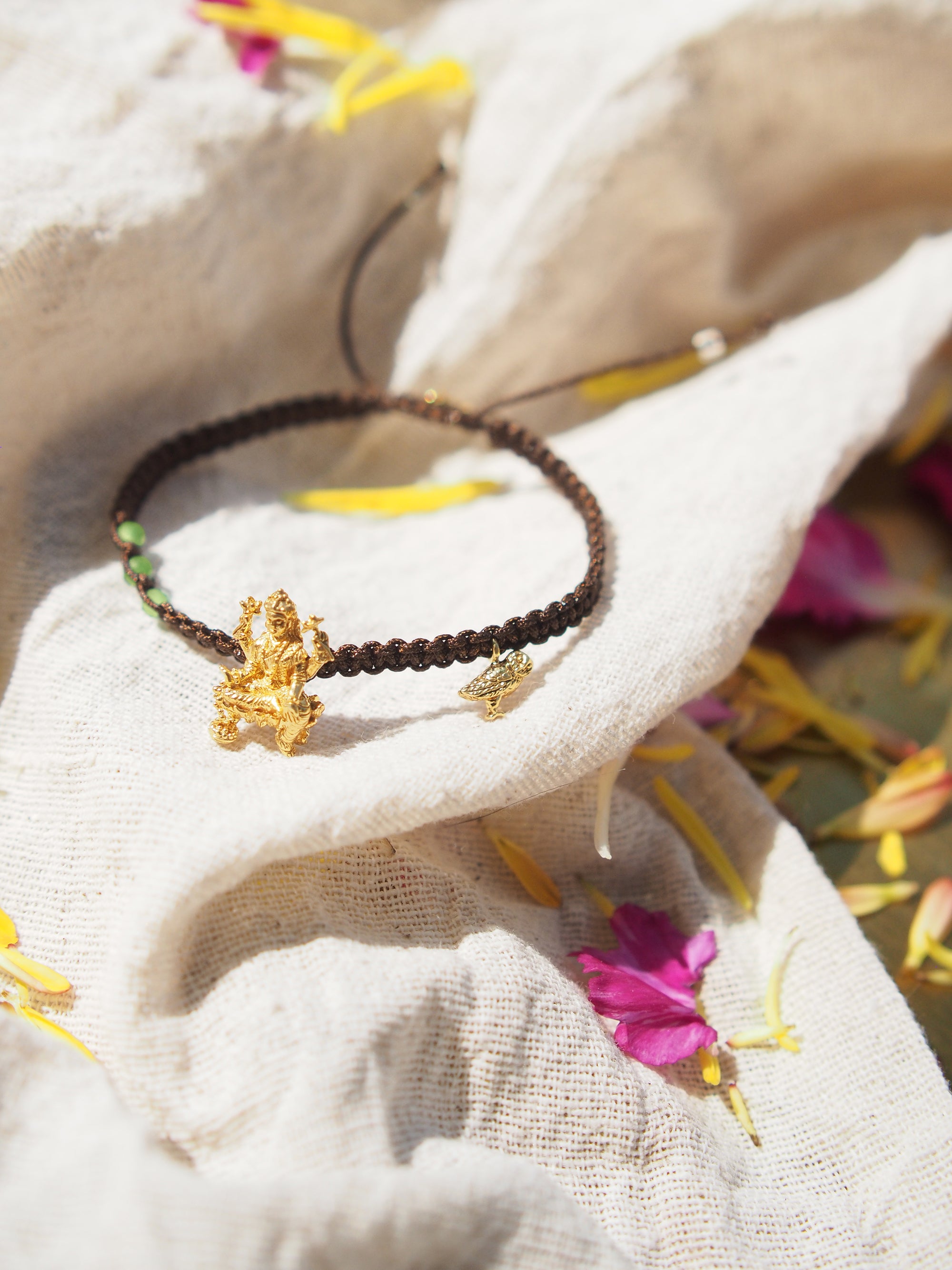 Close-up of a brown bracelet featuring Tridevi Lakshmi and owl charms placed on a fabric among vibrant flowers.