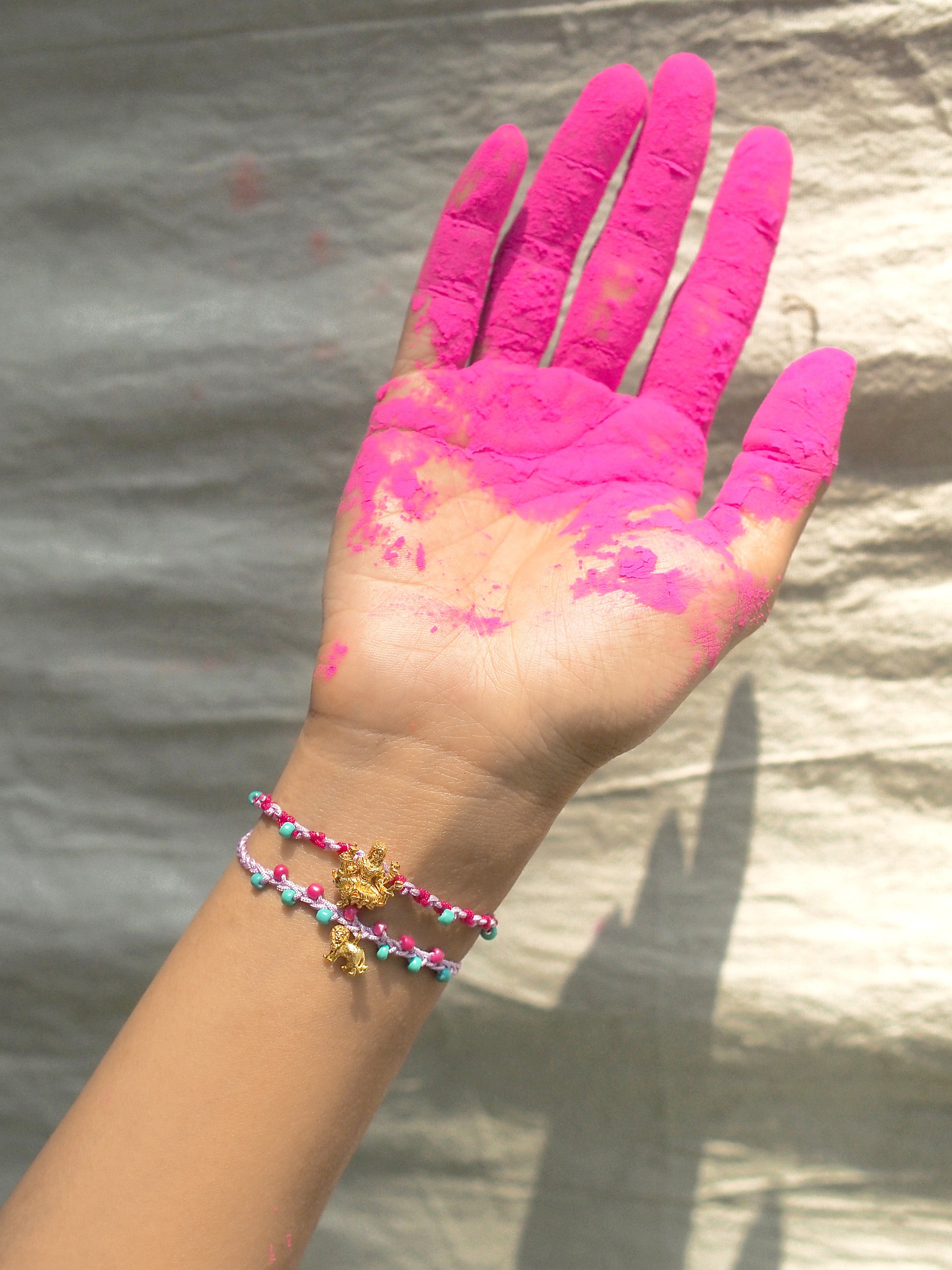A hand with pink powder covering its palm and fingers, while wearing charm bracelet featuring Tridevi Parvati and lion charms.