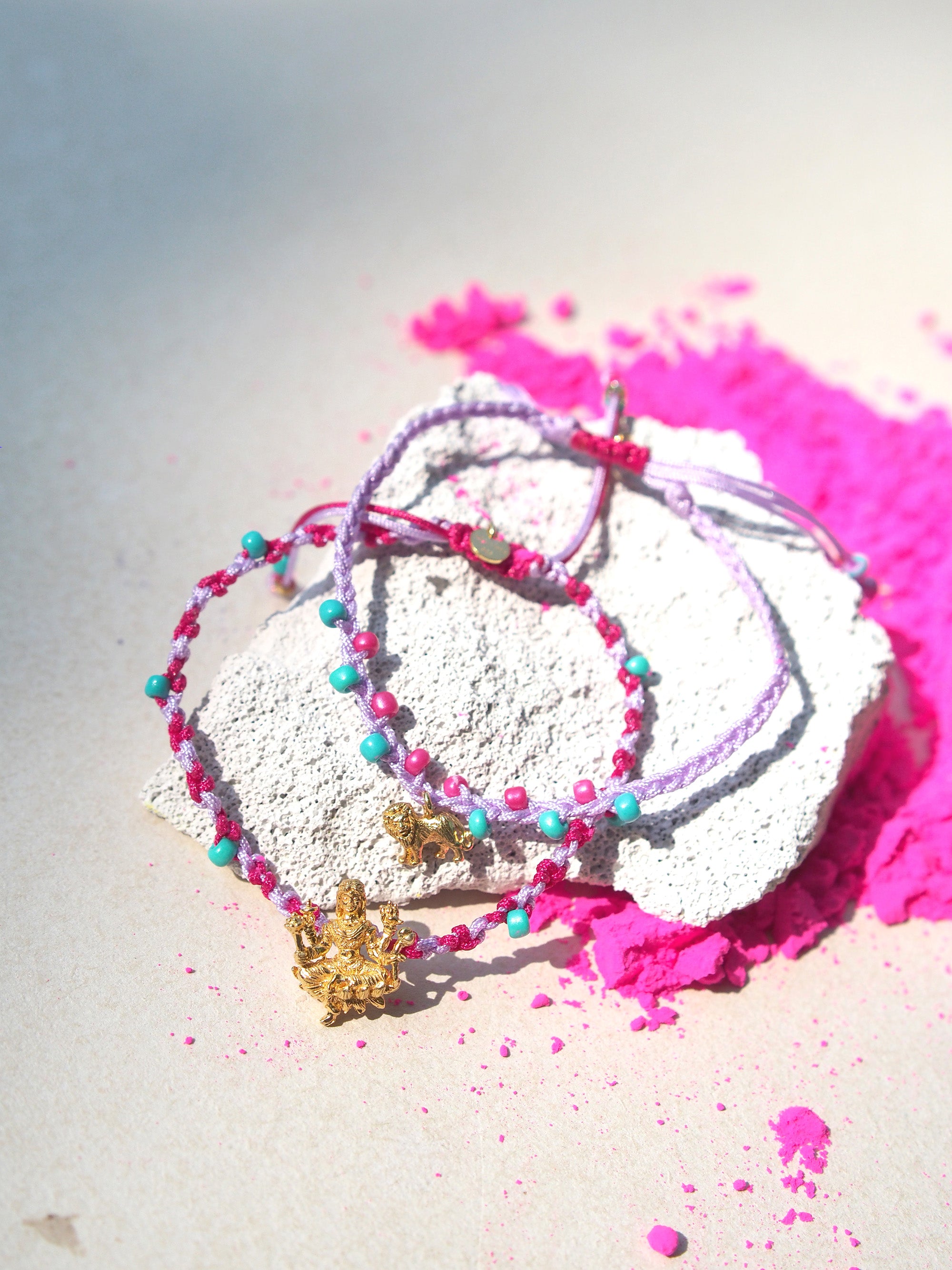 Two bracelets in purple and pink, and turquoise sand beads, featuring a gold Parvati charm and a gold lion charm placed among pink powder.