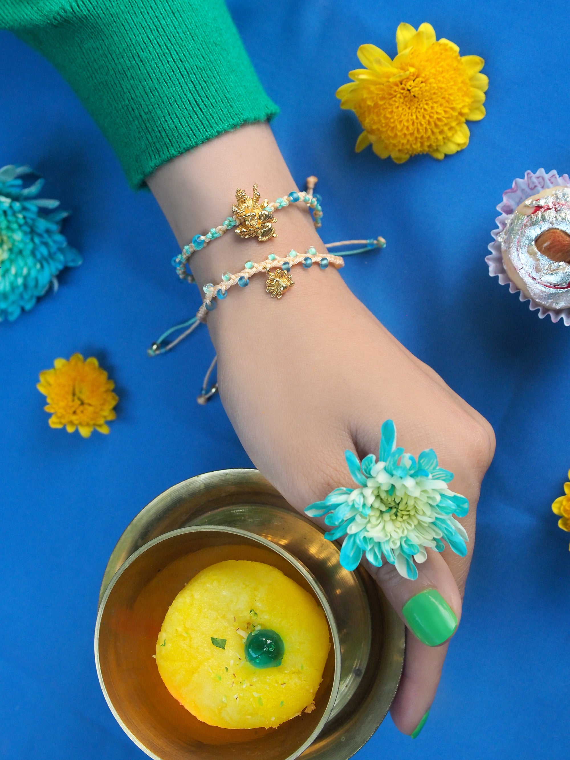 A hand wearing a bracelet set featuring Tridevi Sarasvati and peacock charms, holding a gold bowl with a sweet, surrounded by vibrant flowers.
