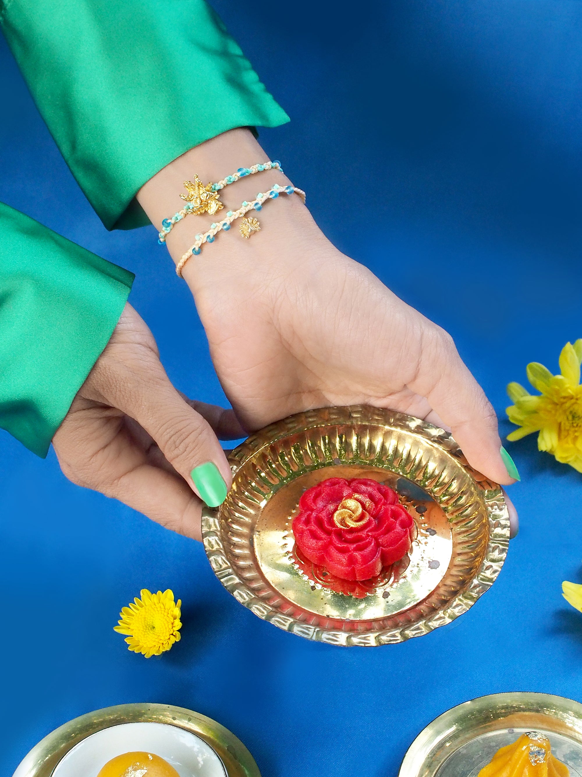A hand wearing a bracelet set featuring Tridevi Sarasvati and peacock charms, holding a gold bowl with a sweet, surrounded by vibrant flowers.