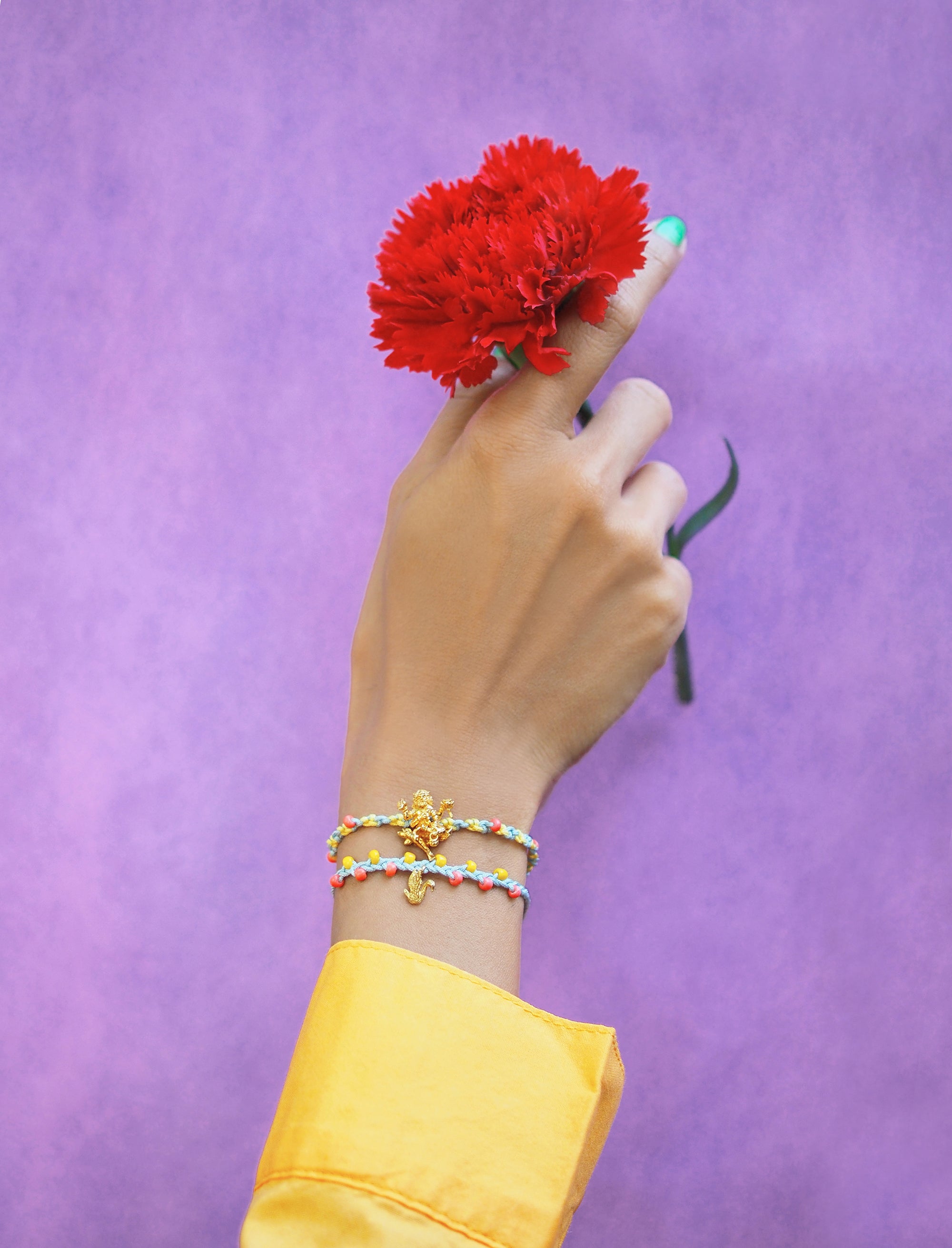 a hand with a Trimurti Brahma charm bracelet set holds a red flower against purple background.