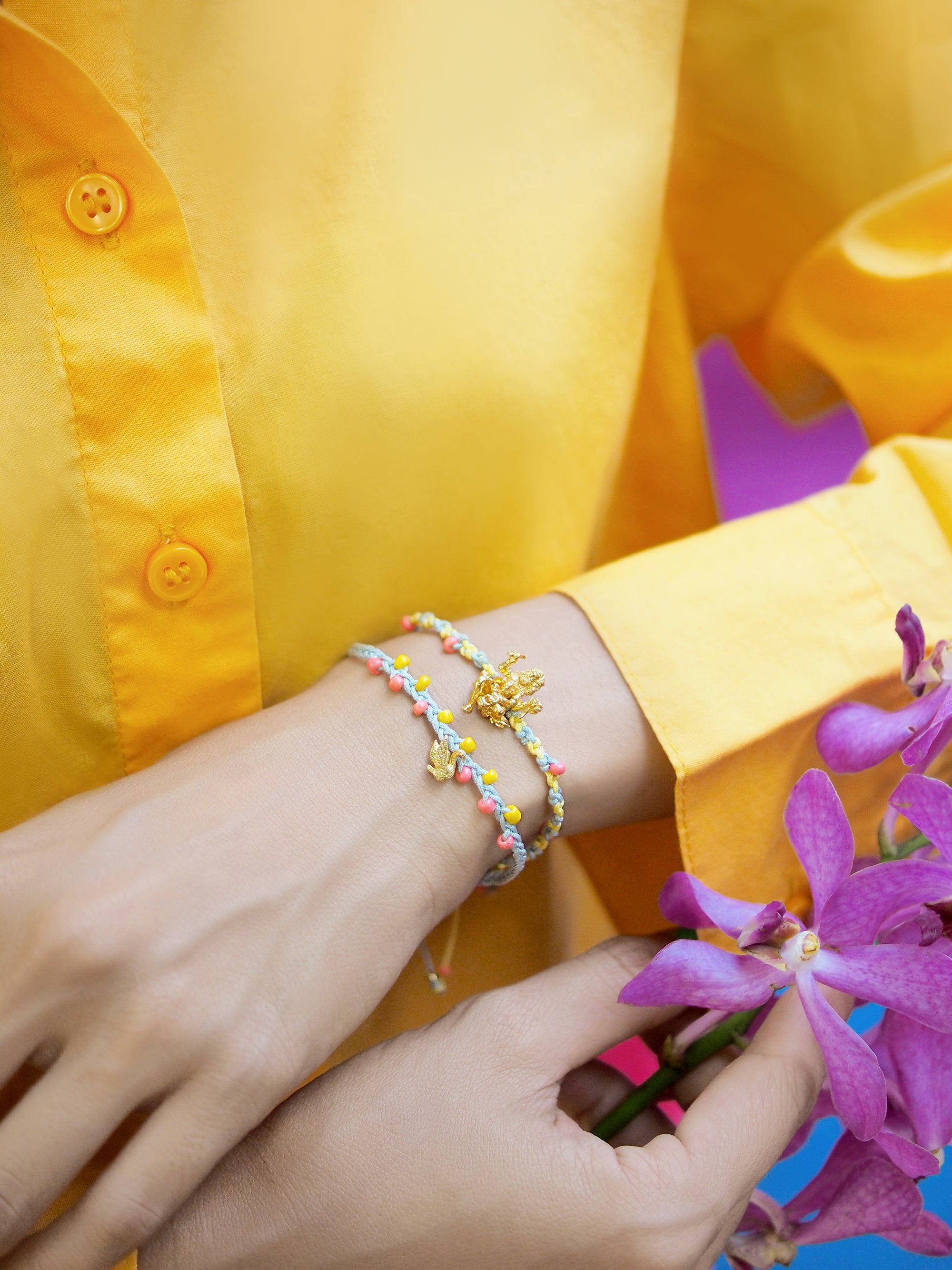 Close-up of person wearing a yellow shirt and a Trimurti Brahma charm bracelet set , while holding purple flowers.