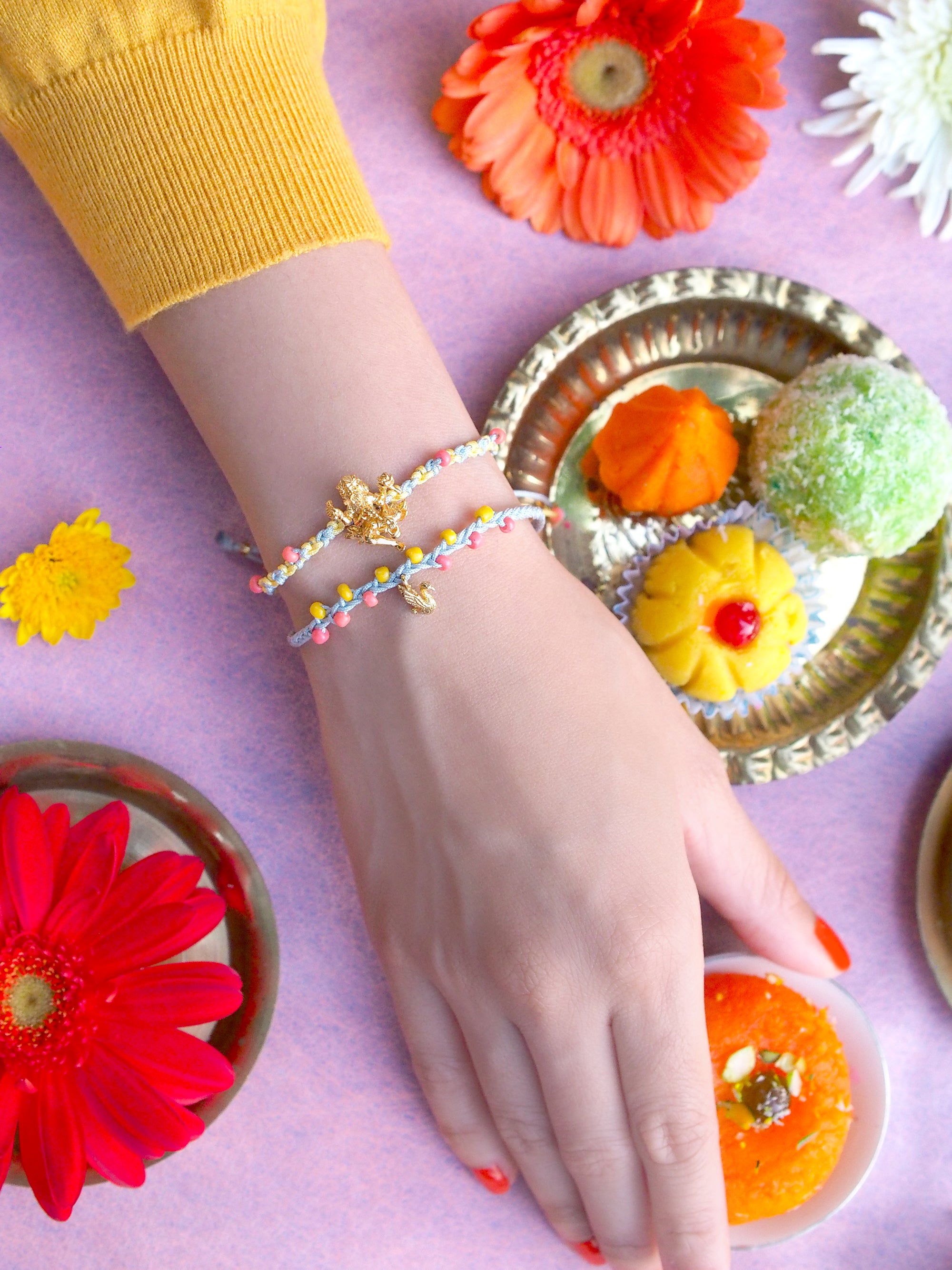 Close-up of a hand with two pastel bracelets featuring a Trimurti Brahma charm and a swan charm, surrounded by festive sweets and flowers