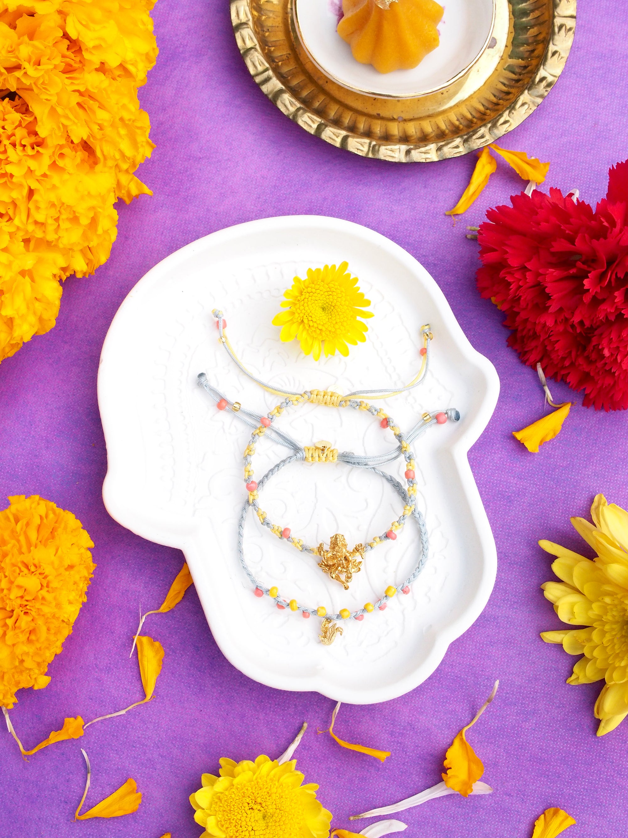 Two bracelets in light grey and  yellow threads, with pastel beads, featuring a gold Trimurti Brahma charm and a gold swan charm surrounded by vibrant flowers.