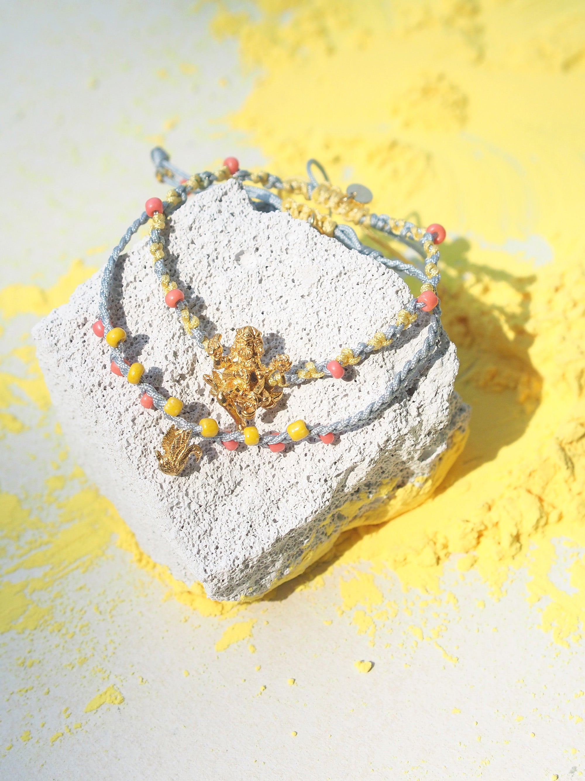 Two bracelets in light grey and yellow with pastel beads, featuring a gold Trimurti Brahma charm and a gold swan charm placed on a textured surface surrounded by yellow powder.