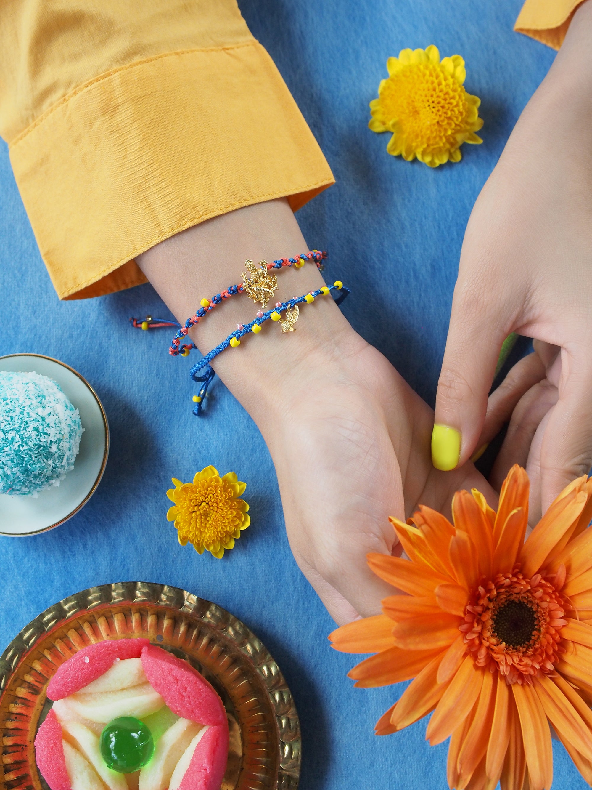 Close-up of a hand with two colorful bracelets featuring a Trimurti Vishnu charm and a garuda charm, surrounded by festive sweets and flowers