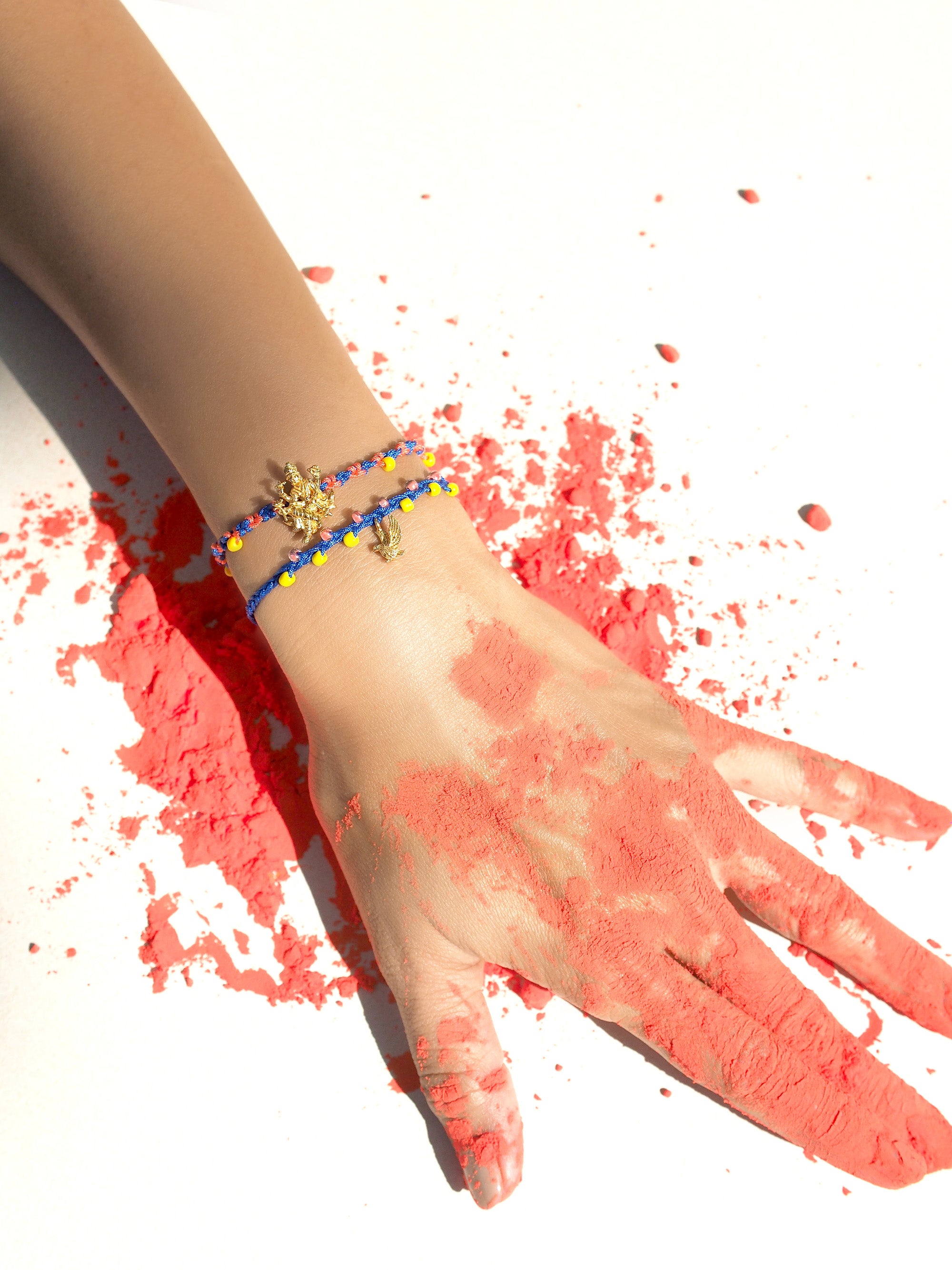A hand with red powder covering its fingers, while wearing charm bracelet featuring Trimurti Vishnu and garuda charms.
