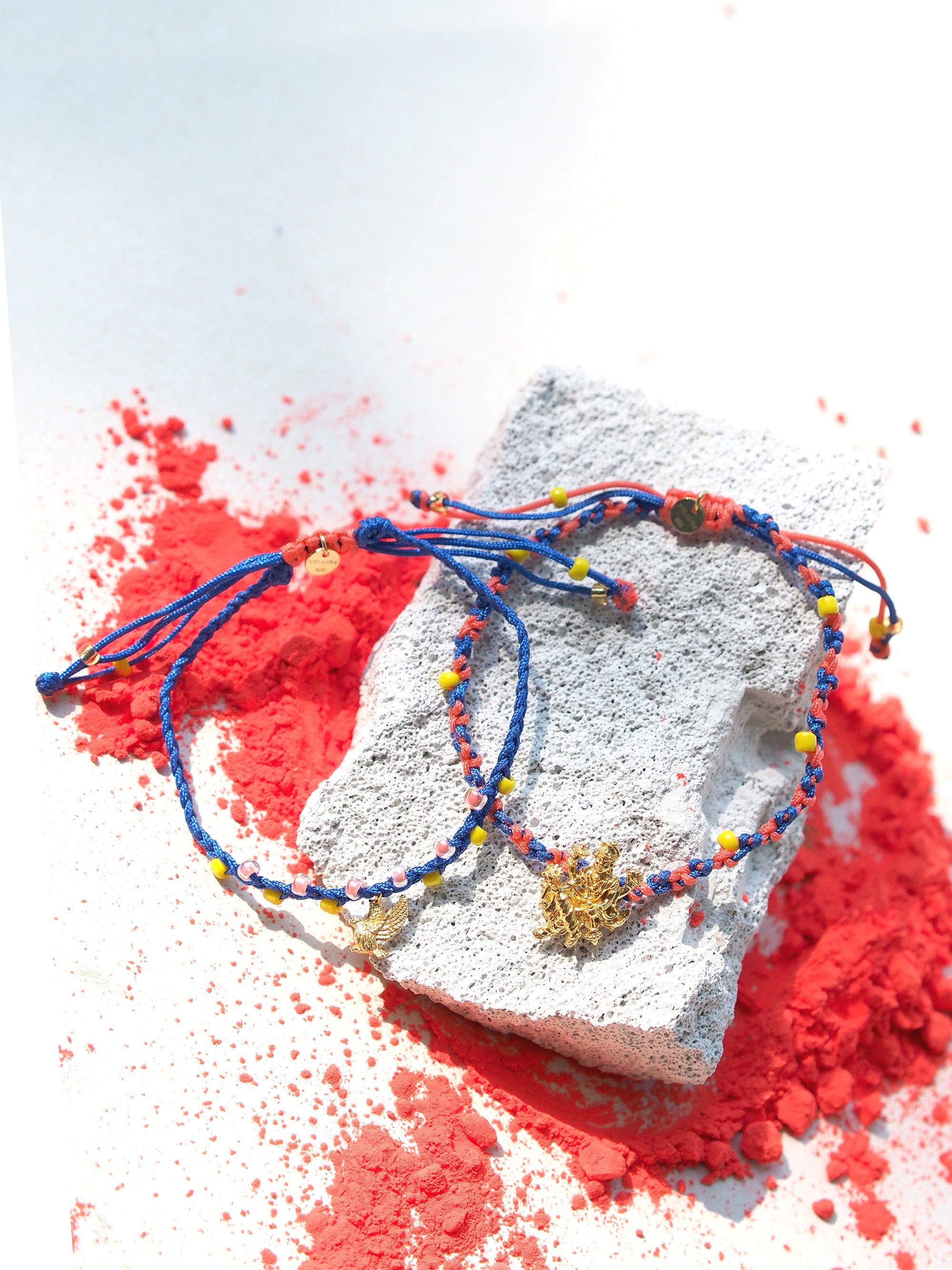 A colorful bracelet set featuring a gold Trimurti Vishnu charm and a gold garuda charm placed on a textured surface among red powder.