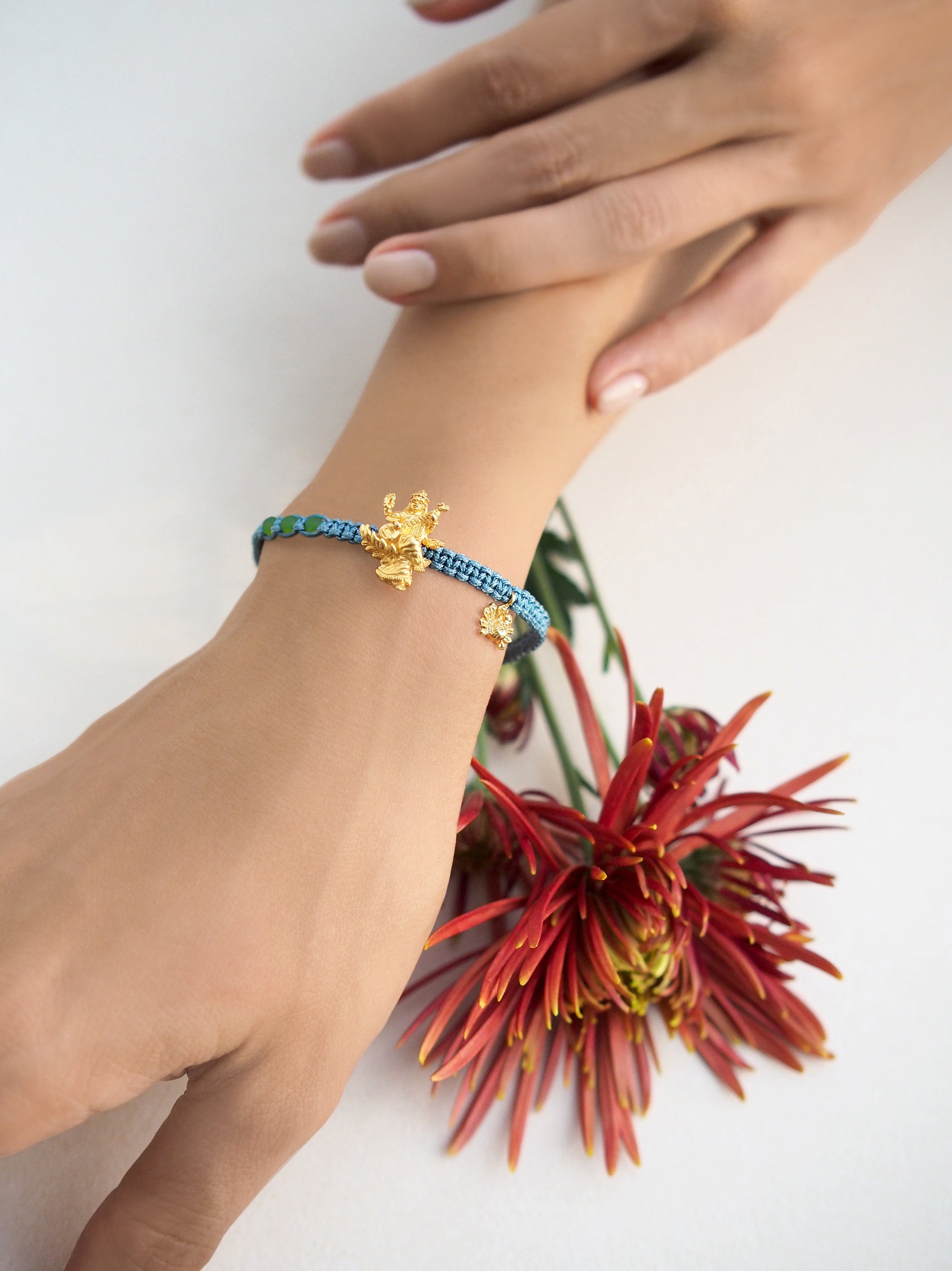 Close-up of a green bracelet adorned with a gold Tridevi Sarasvati charm worn on wrist against red flowers in the background.