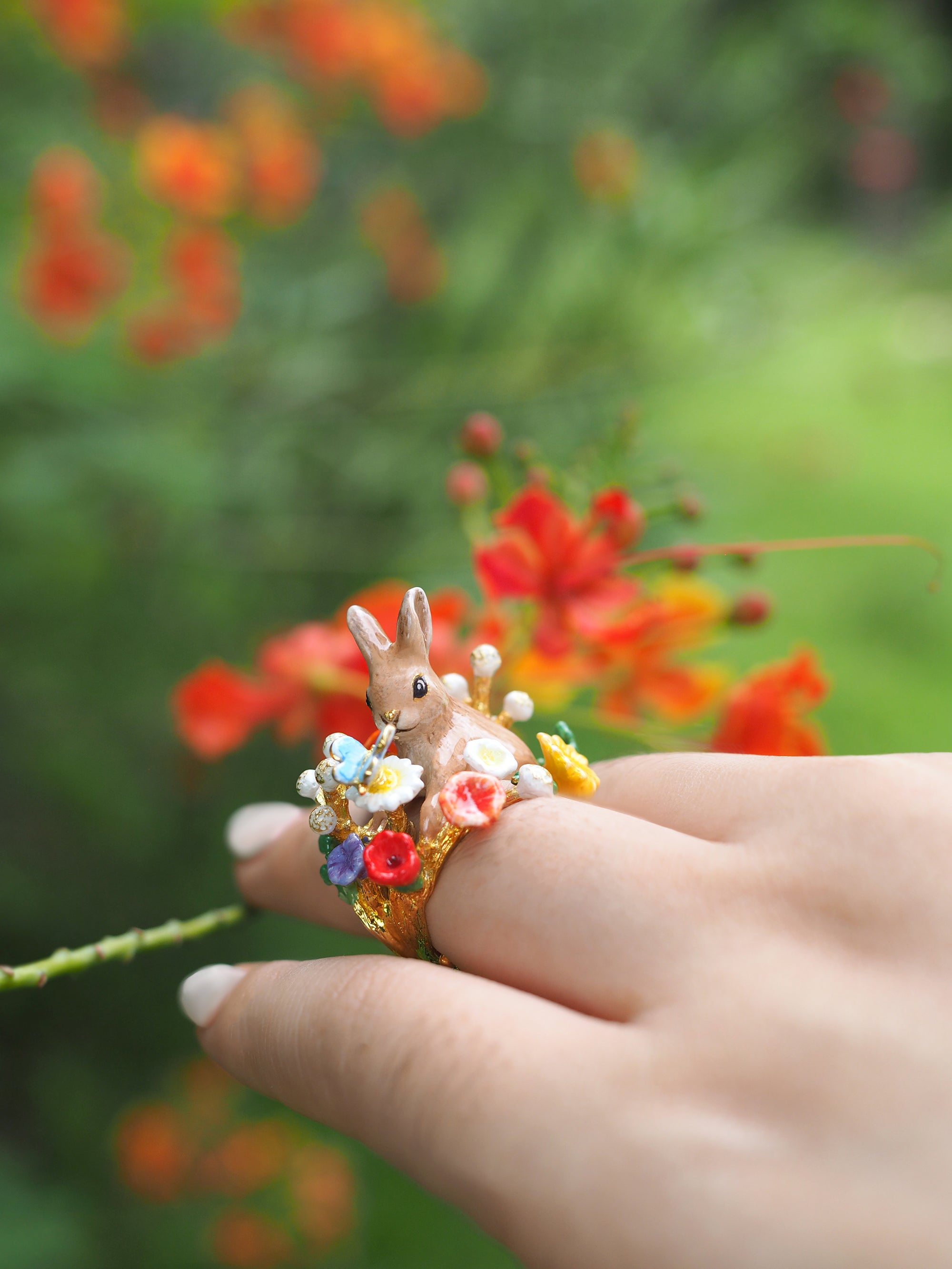 Close-up of a hand with a ring featuring a rabbit surrounded by colorful flowers and a blue butterfly, with a natural setting.