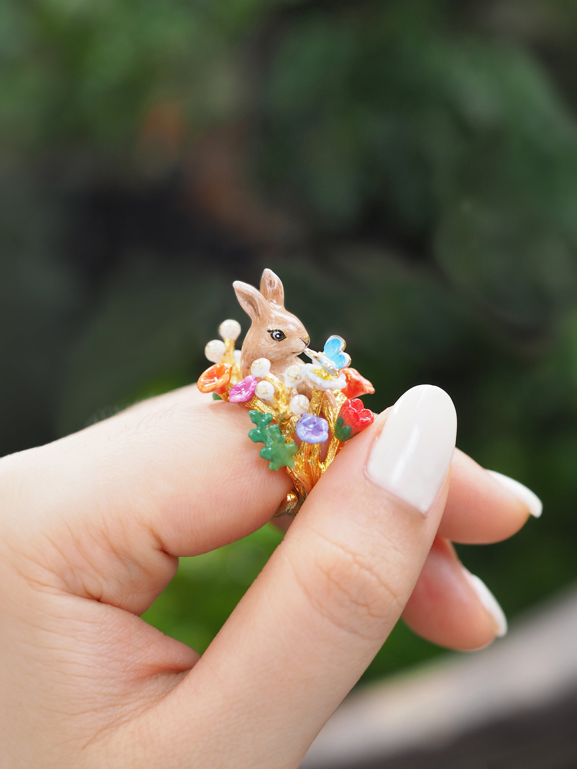 Close-up of a hand showcasing a ring featuring a rabbit surrounded by colorful flowers and a blue butterfly in a greenery setting.