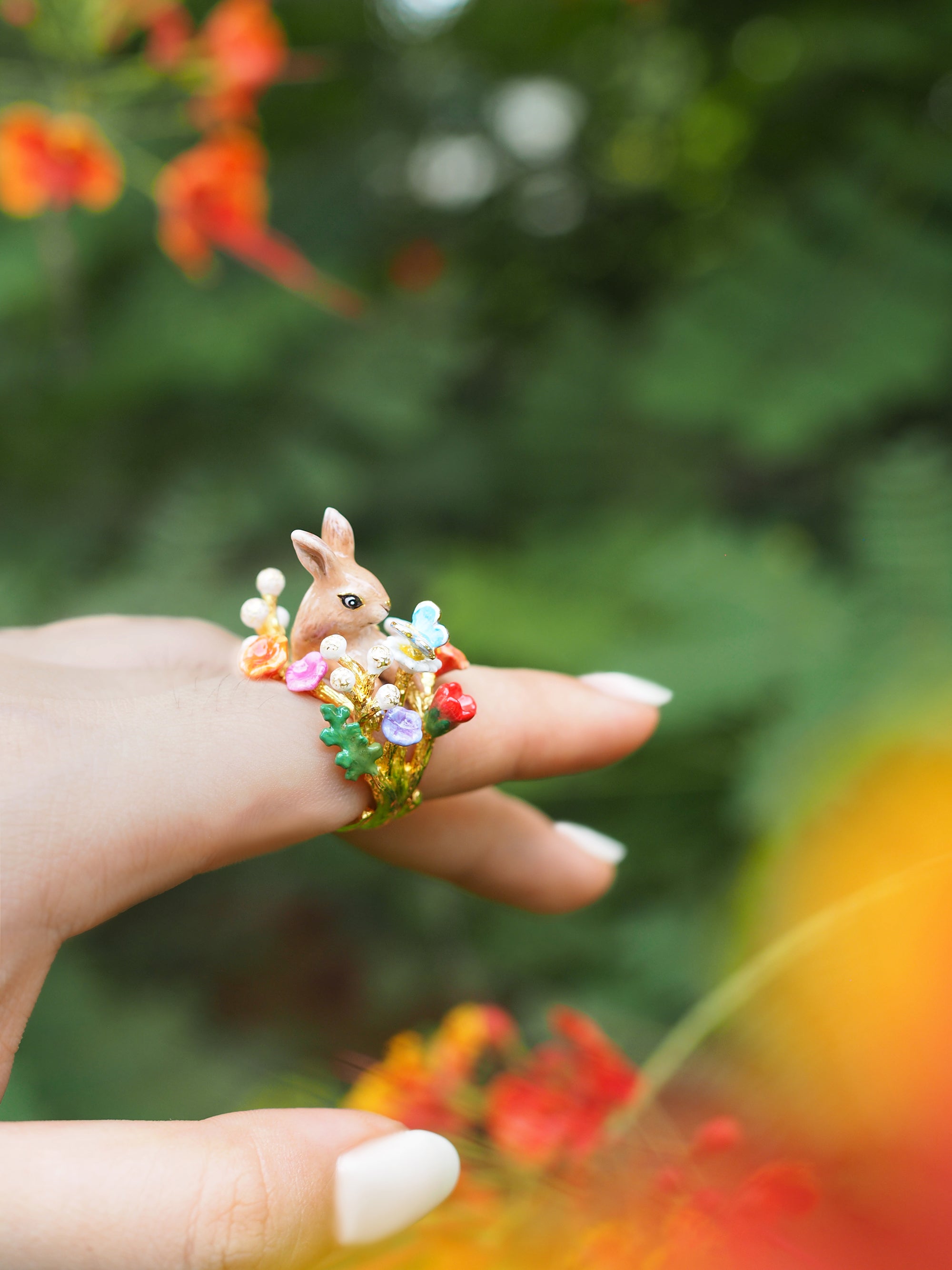 Close-up of a hand wearing a ring featuring a rabbit surrounded by colorful flowers and a blue butterfly, with natural background.