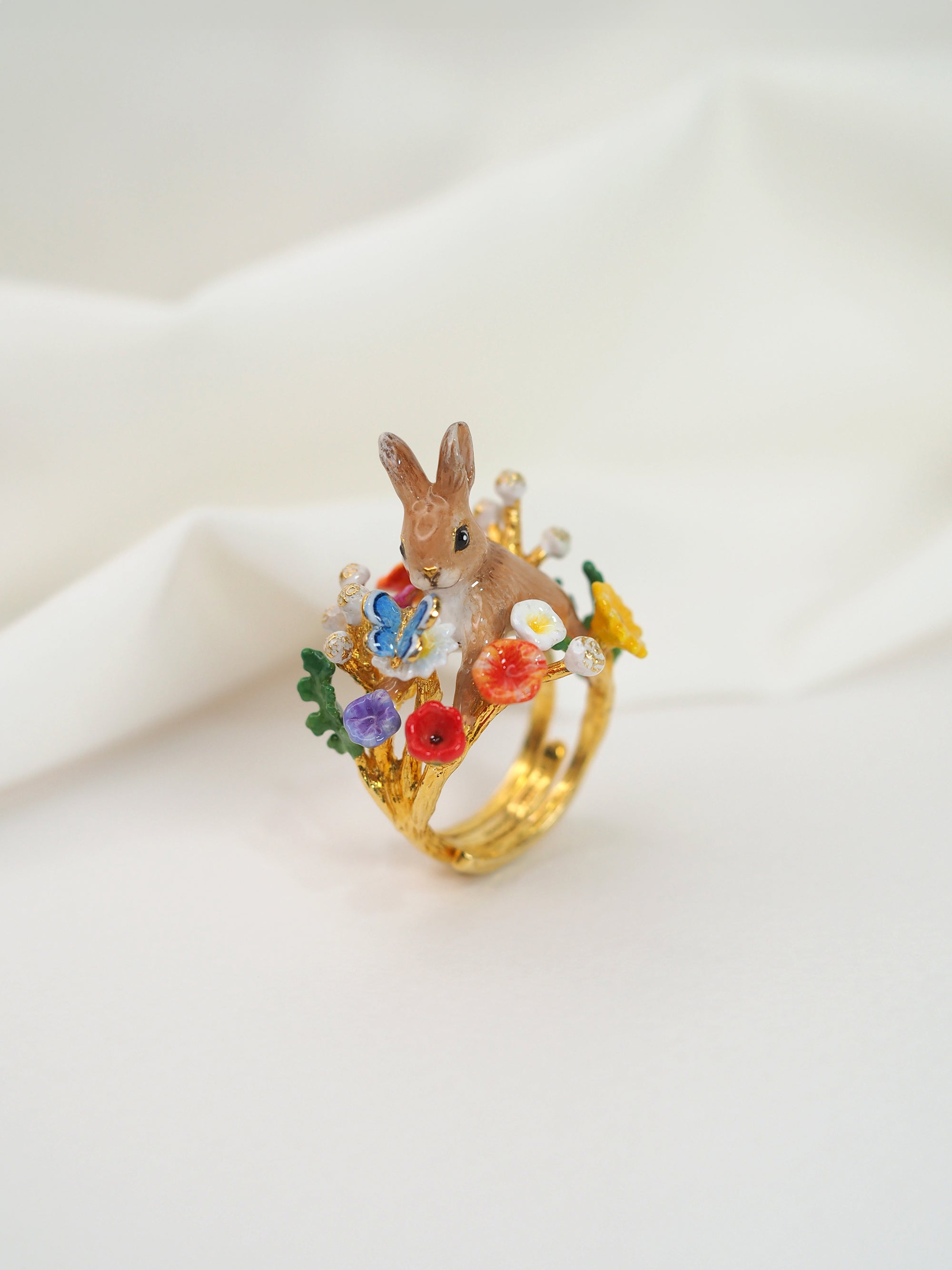 A close-up of an adorable ring featuring a rabbit surrounded by colorful flowers placed on a white fabric.