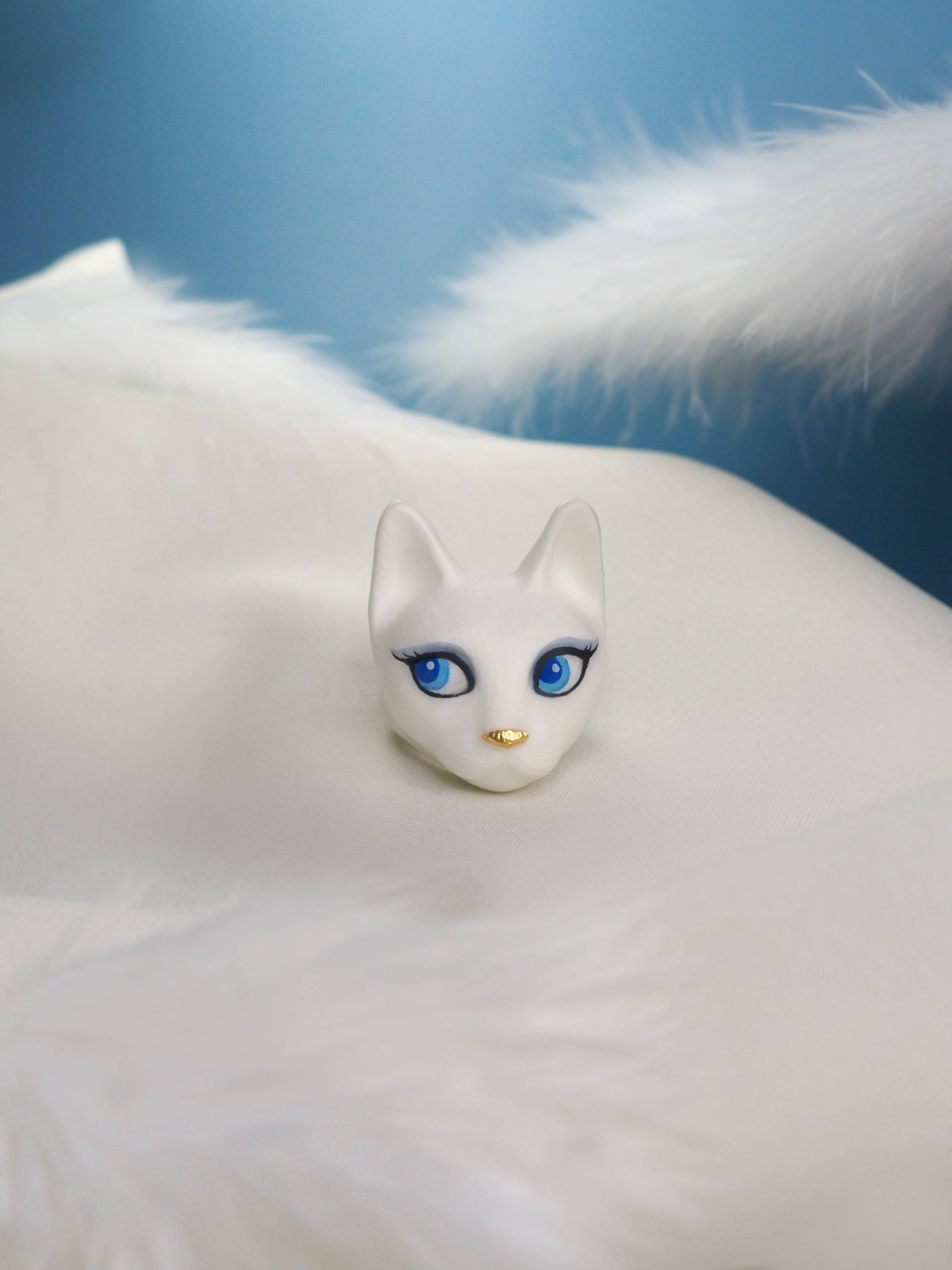 A hand-painted ring showcasing a white cat with captivating blue eyes placed on white fabric with feather elements.