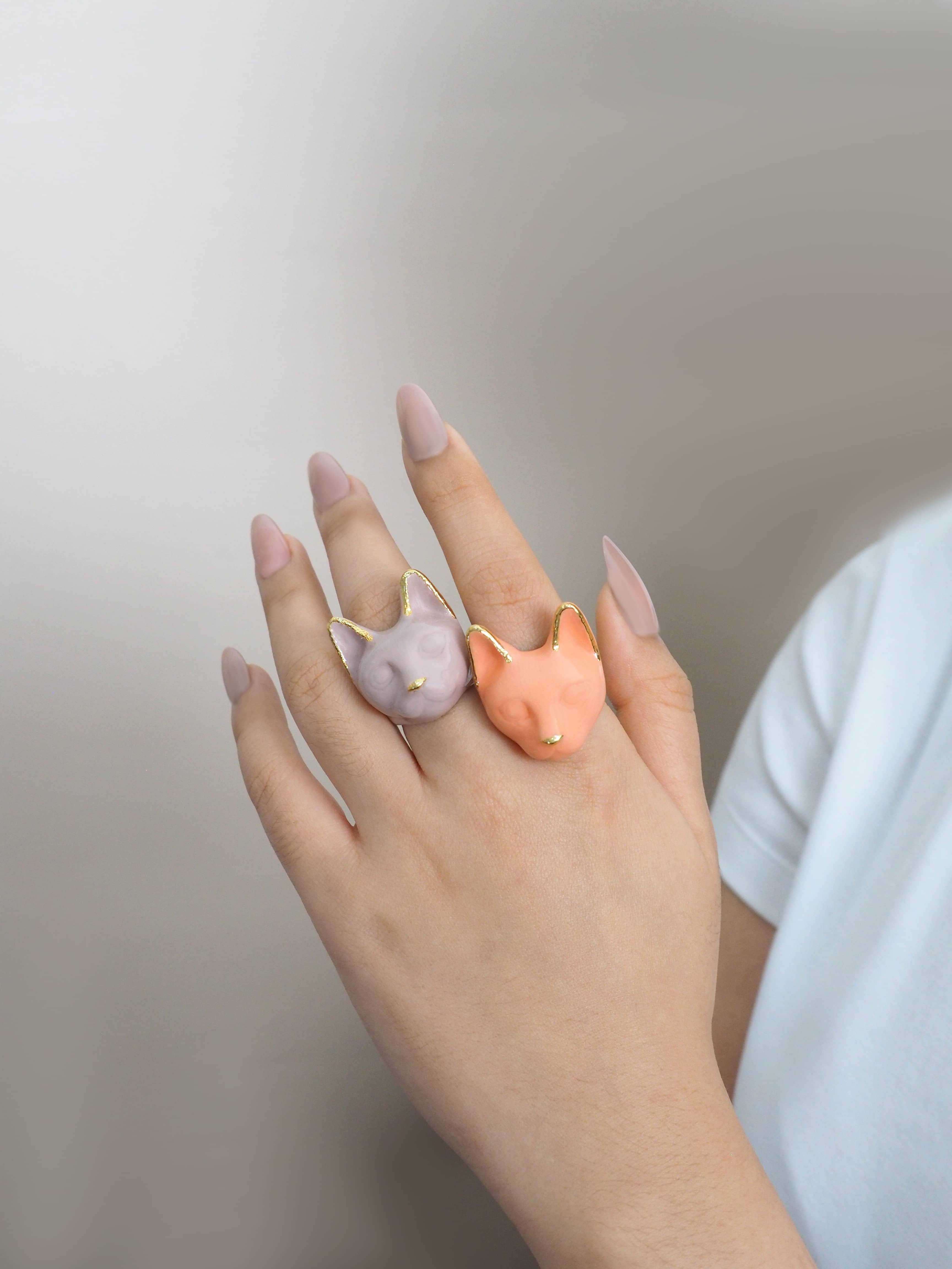 A hand wearing two cat rings: orange and dusty pink, perfect for art collectors.