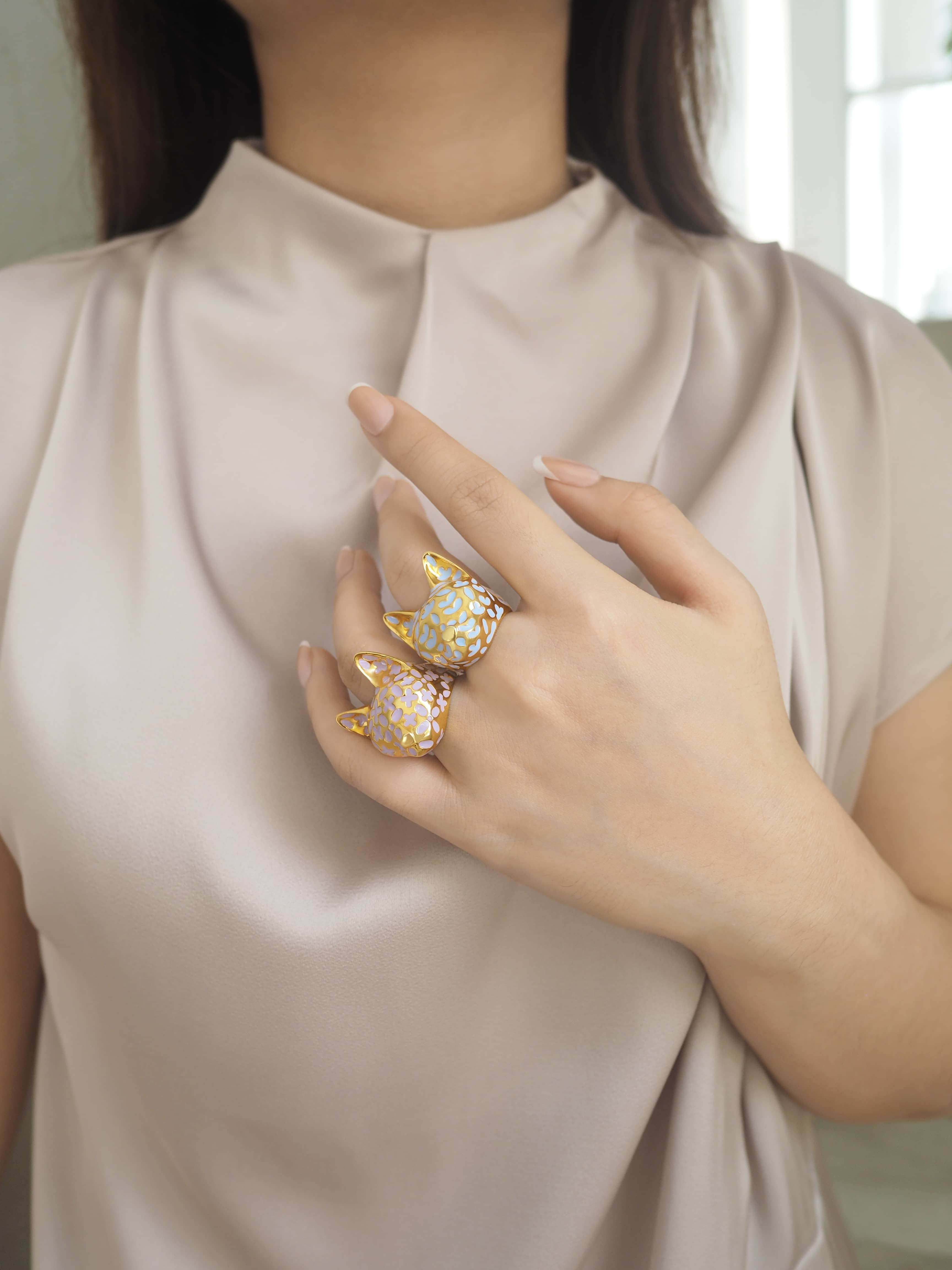 A close-up of a hand adorned with two gold cat rings with purple flower patterns, inspired by lilac and lavender.