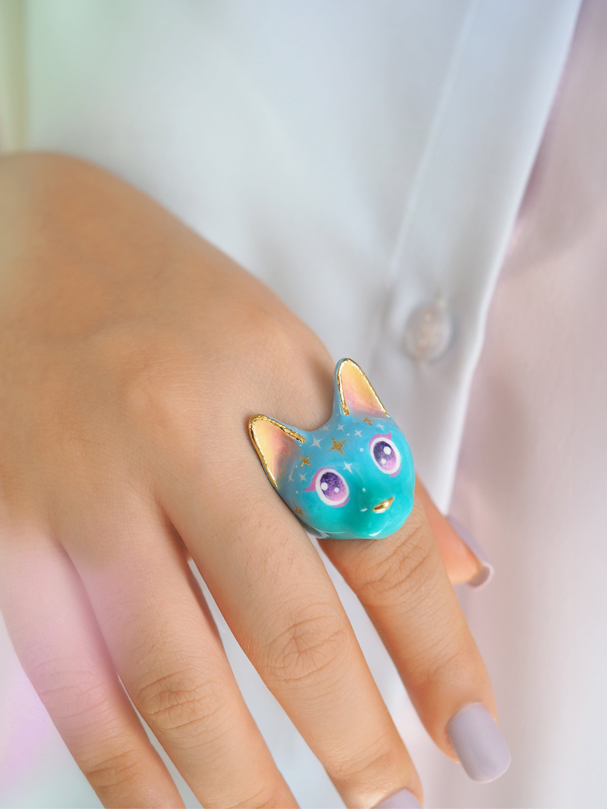 A close-up of a hand with a cat ring in a soft blue color featuring sparkling purple eyes and star-like patterns on its forehead.