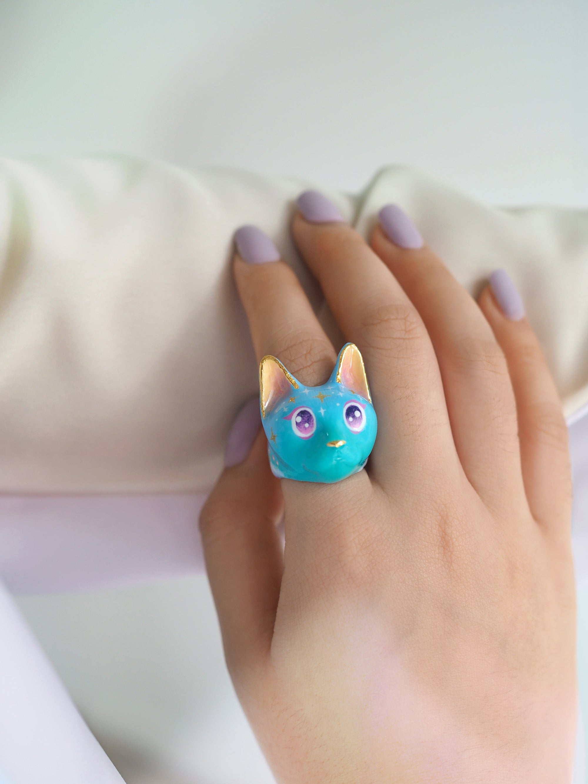 A close-up of a whimsical ring with a cat's face design in a soft blue color featuring yellow ears and large, sparkling purple eyes and star-like patterns on its forehead