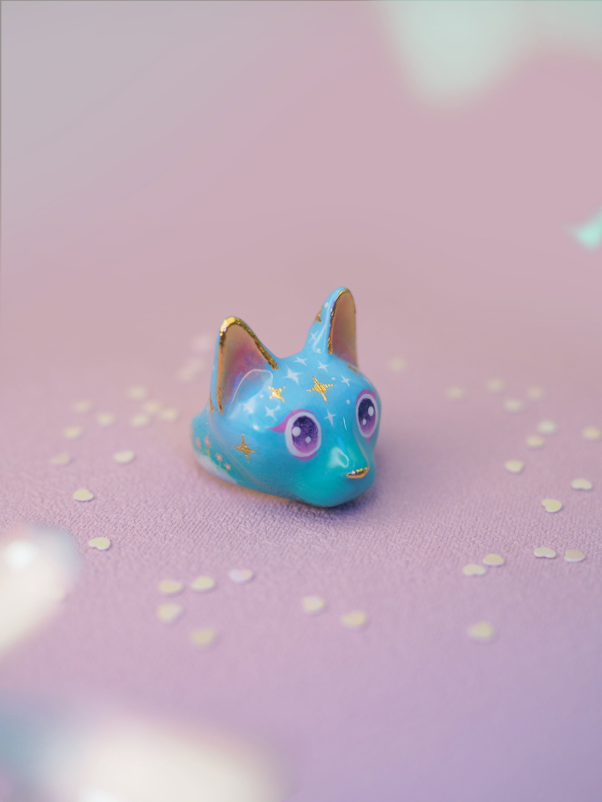 Hand-painted cat ring featuring a blue face, yellow ears, sparkling purple eyes, and starry patterns placed on a light pink background.