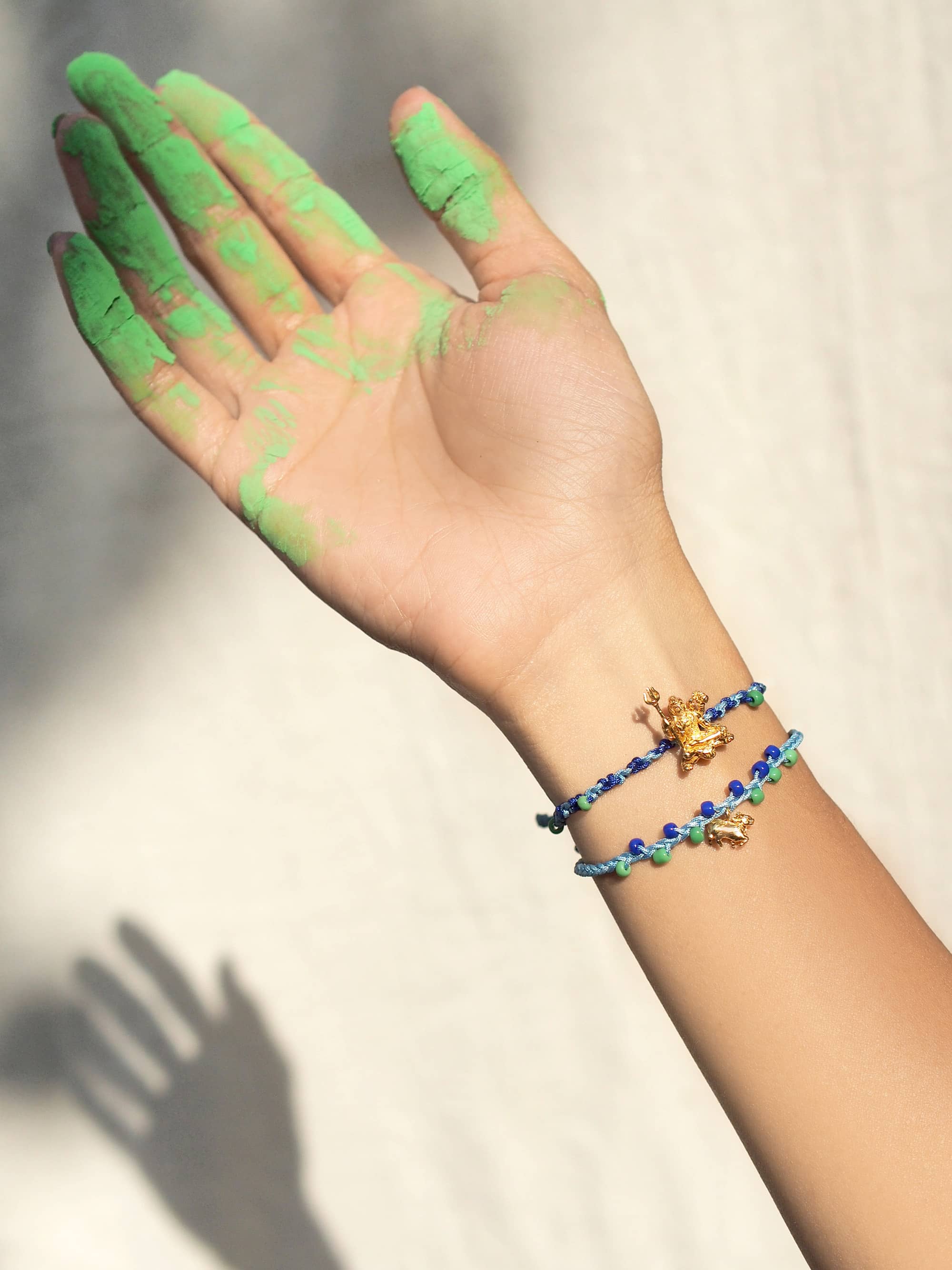 A hand with green powder covering its fingers, while wearing a colorful bracelet featuring Trimurti Shiva and his Nandi charms.