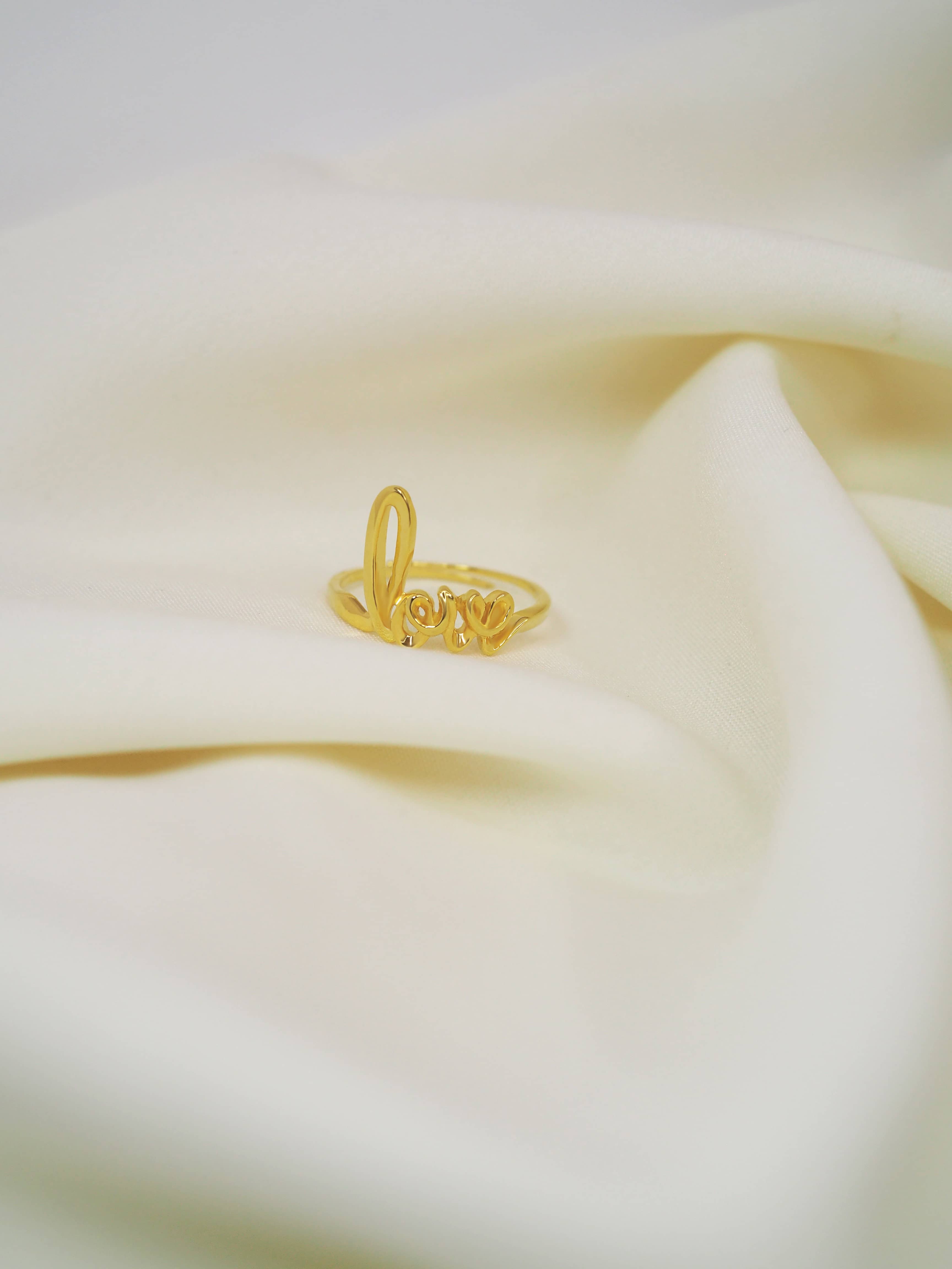 Beautiful designed gold ring featuring the word 'love' in a handwritten style placed in white fabric.