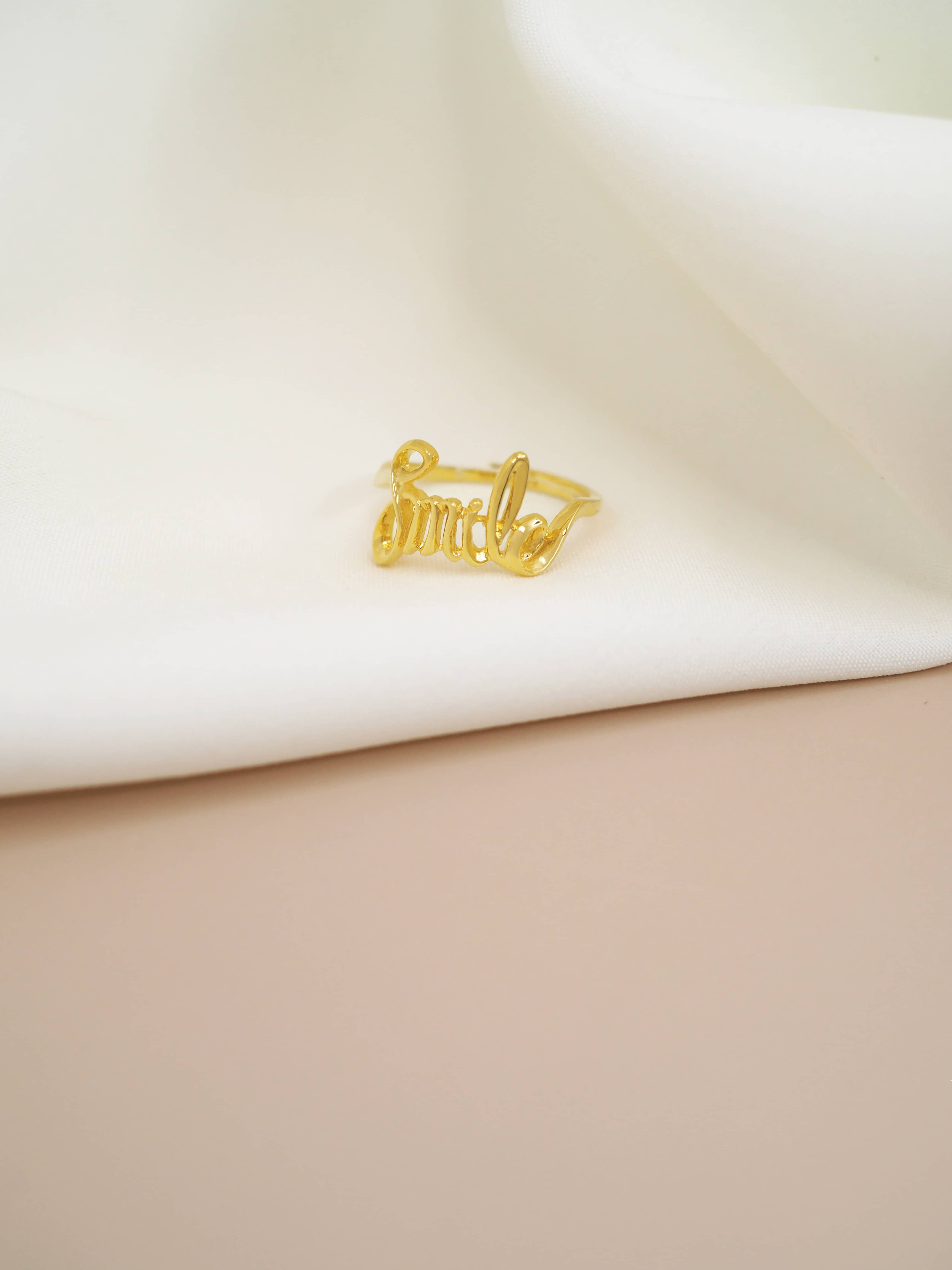 Stylish gold ring featuring the word 'Smile' in a handwritten style placed on white fabric.