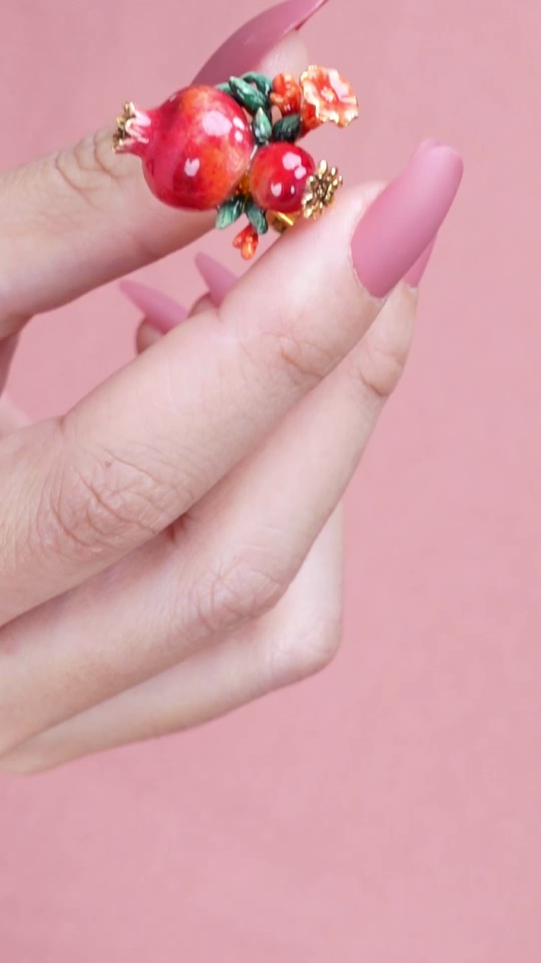 A person wearing a charming ring featuring two pomegranates, green leaves, and small red flowers, ideal for summer fashion.