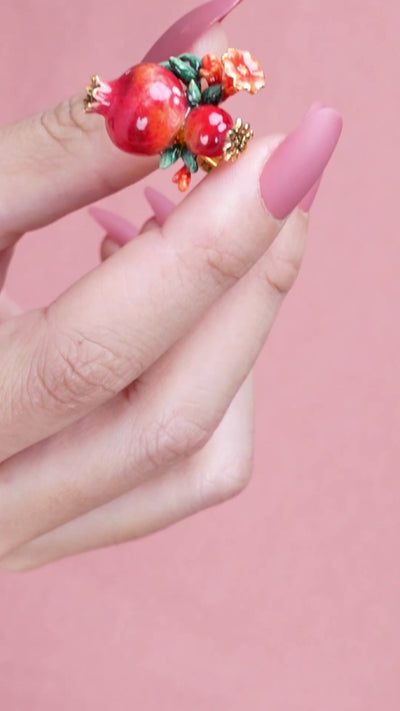 A person wearing a charming ring featuring two pomegranates, green leaves, and small red flowers, ideal for summer fashion.