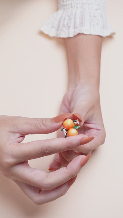 A hand wearing a handmade ring featuring two oranges, green leaves, and small white flowers, in a summer outfit.