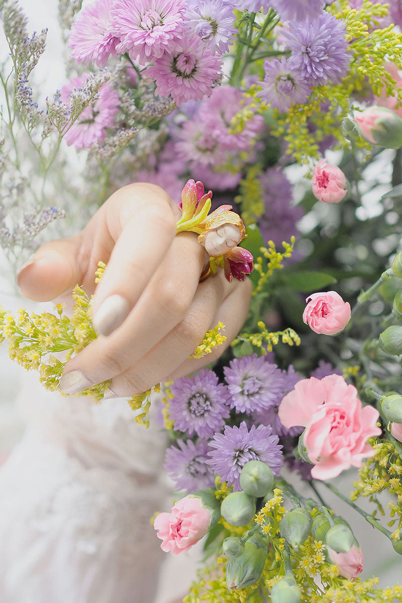 A close-up of a hand wearing a beautiful nymph ring surrounded by colorful flowers.