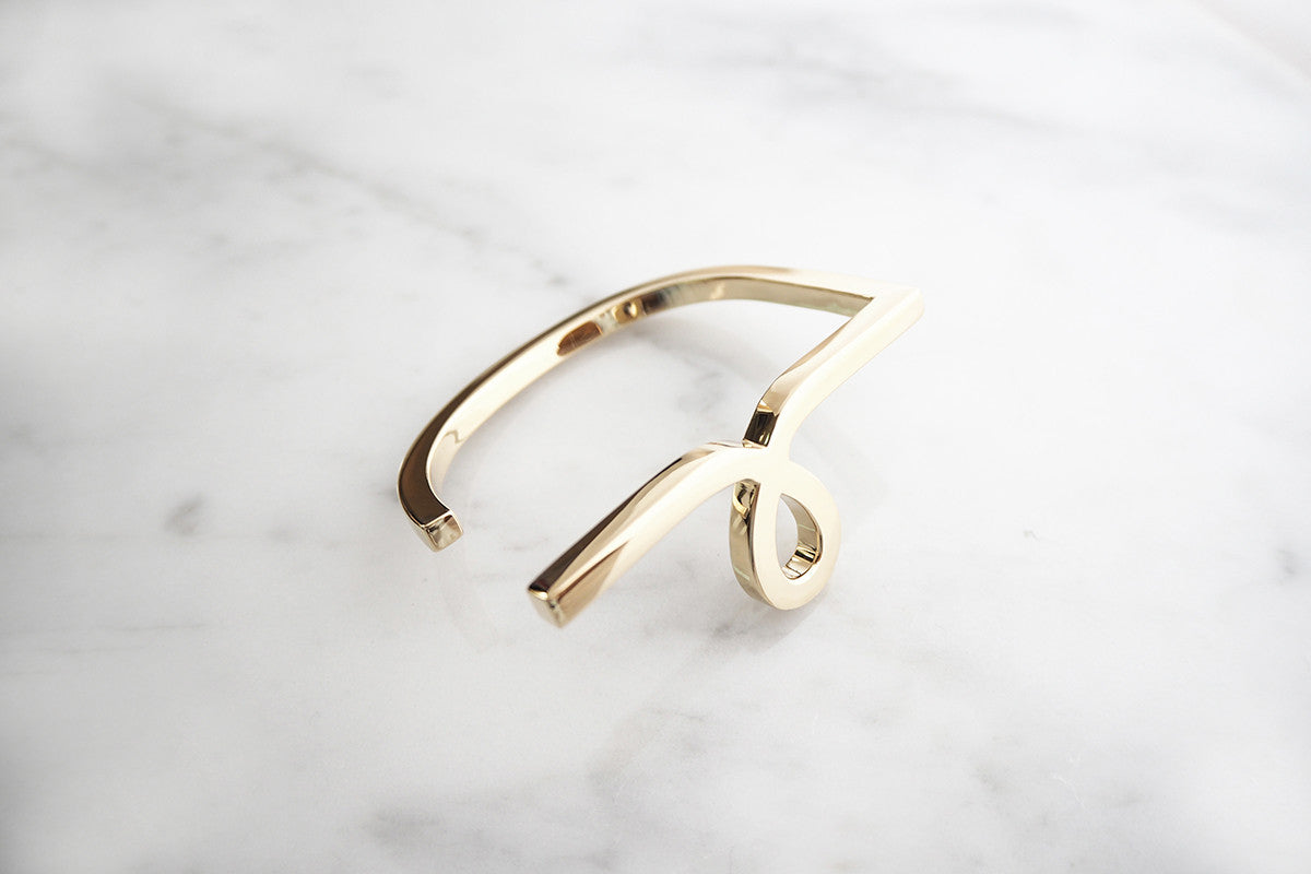Gold bangle featuring a unique script-style small "L" design placed on a marble background.