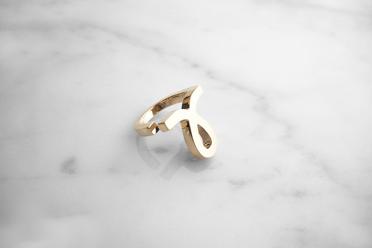 Gold ring featuring a unique script-style small "L" design placed on a marble background.