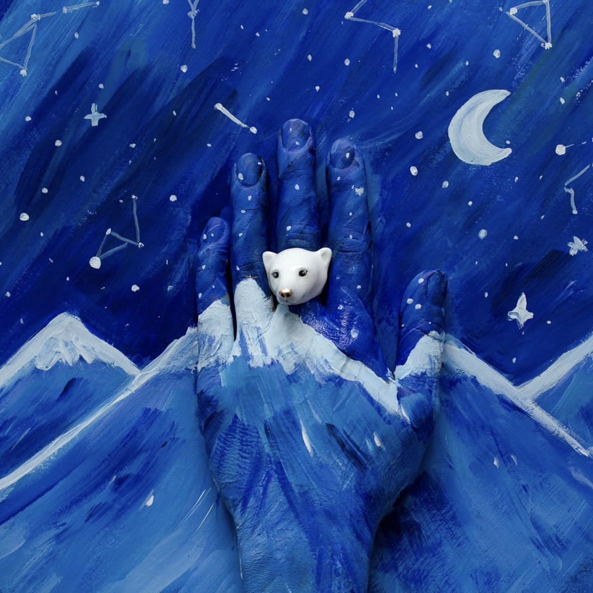 A blue-painted hand wearing a white polar bear ring against a starry night sky background with painted constellations, a crescent moon, and mountains.