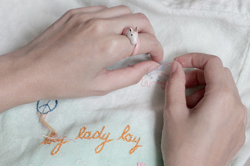 A hand wearing a cute unicorn ring stitches a pink design onto fabric.
