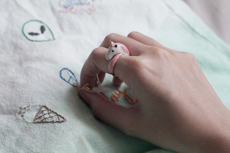 A hand wearing a cute unicorn ring stitches a pink design onto fabric.