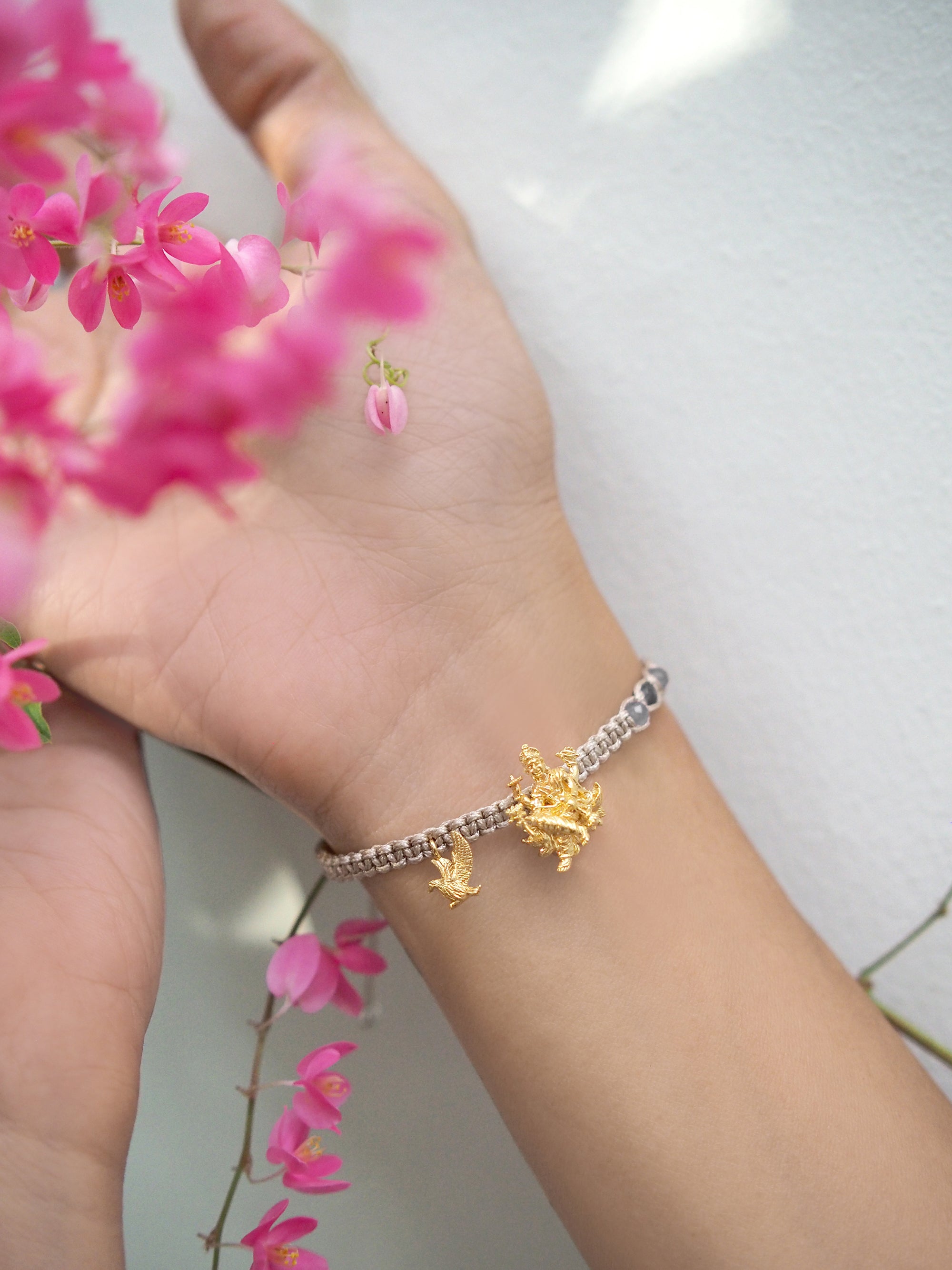 A hand wearing an elegant bracelet featuring Trimurti Vishnu and Garuda charms surrounded by pink flowers.