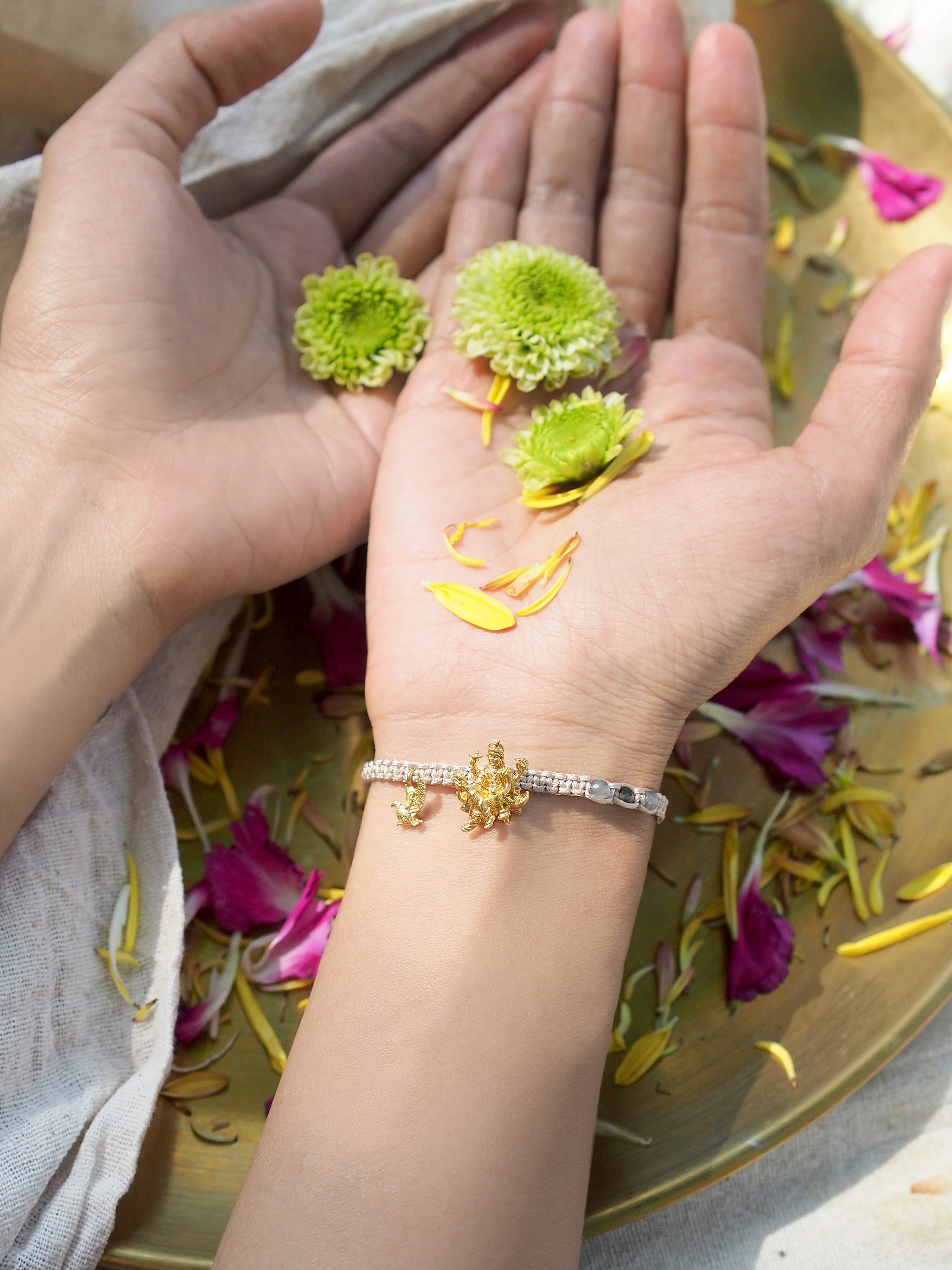 A hand with a Vishnu bracelet holds green flowers and petals in natural setting