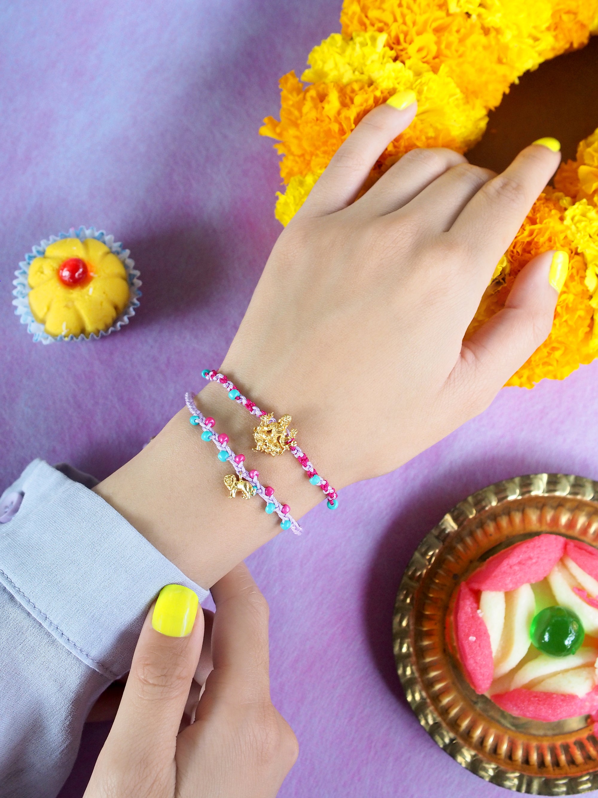 Close-up of a hand with two colorful bracelets featuring a Tridevi Parvati charm and a lion charm, surrounded by festive sweets and flowers