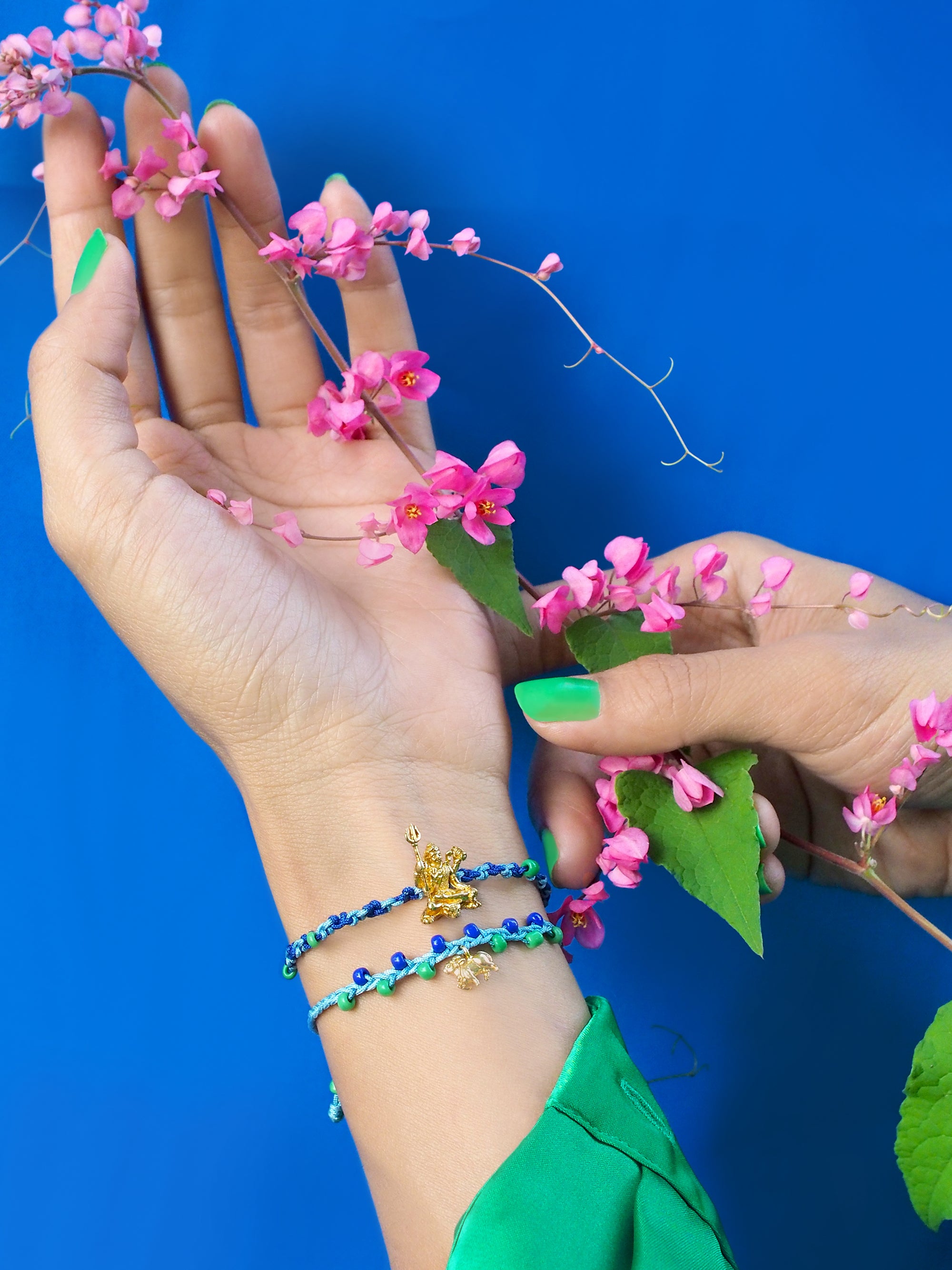 Colorful Trimurti Shiva bracelets adorn a hand amidst pink flowers against a blue background.