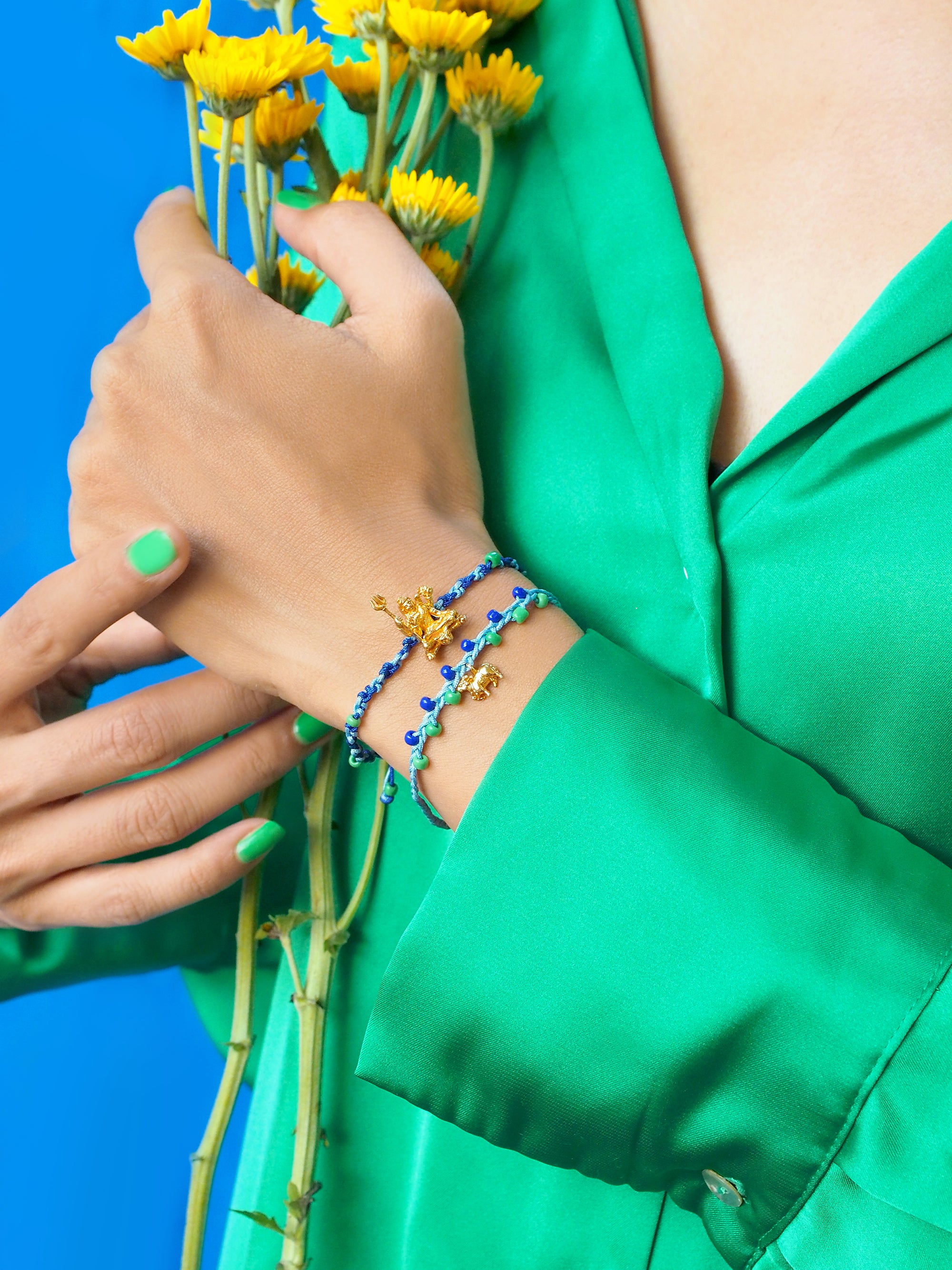 A person wearing a green shirt and a colorful Trimurti Shiva bracelet set on a wrist while holding flowers against a blue background.