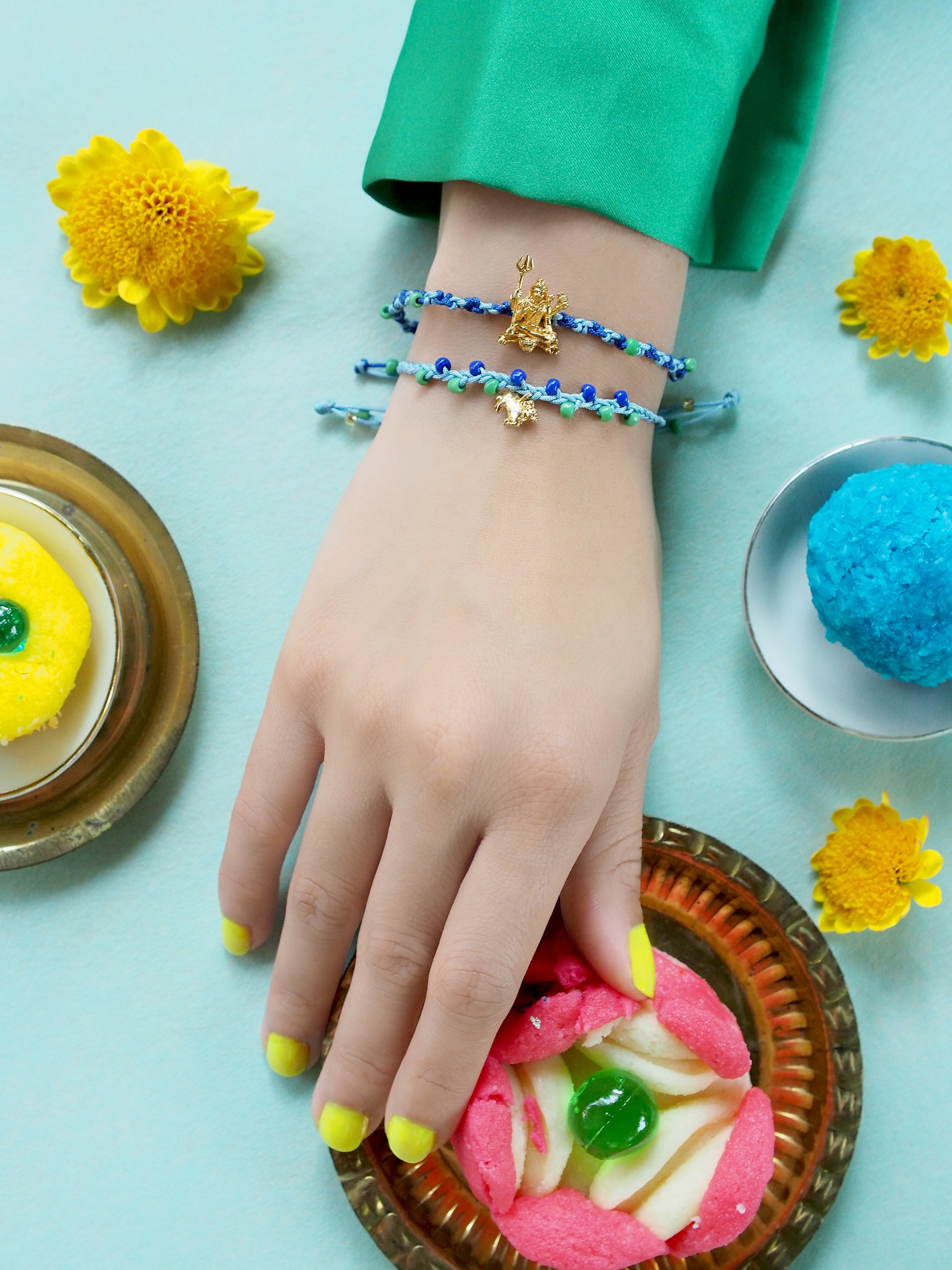 Close-up of a hand with two bracelets featuring a Trimurti Shiva charm and a Nandi charm, surrounded by festive sweets and flowers